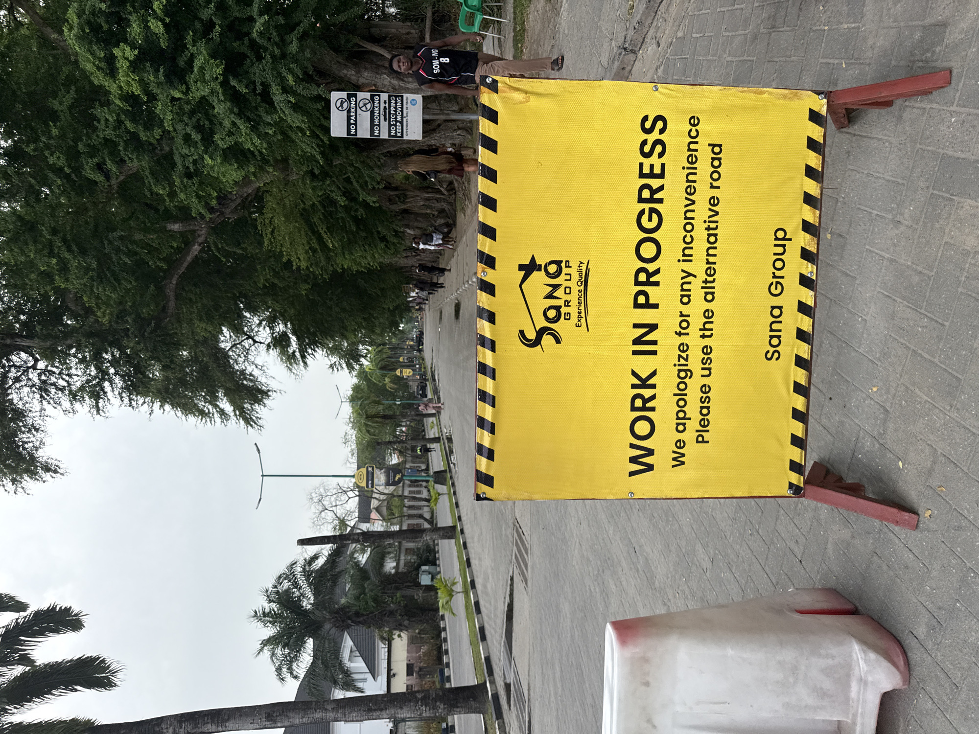 The image displays a street scene in Lekki, Nigeria, featuring a large yellow banner indicating "WORK IN PROGRESS" prominently in the foreground on the right. The banner reads: "WORK IN PROGRESS", "We apologize for any inconvenience Please use the alternative road", and includes the "Sana Group Experience Quality" logo and company name. It is supported by a red, wooden-like stand.
To the left of the banner, a man wearing a black and white shirt with "SOM-NG 8" on it stands near a large tree with dense green foliage. Behind him, a black and white sign is mounted on a post, detailing traffic regulations: "NO PARKING", "NO HONKING", and "NO STOPPING KEEP MOVING OFFENDERS WILL BE FINED".
The scene is an outdoor setting, likely a street or pathway, with a paved surface. Lush green trees, including several palm trees, line the thoroughfare, extending into the background. Buildings are visible further down the street. The sky appears overcast, suggesting a daytime shot. A white and red barrier or cone is partially visible at the bottom right, next to the banner. The image captures a moment of ongoing activity or construction, necessitating traffic adjustments.