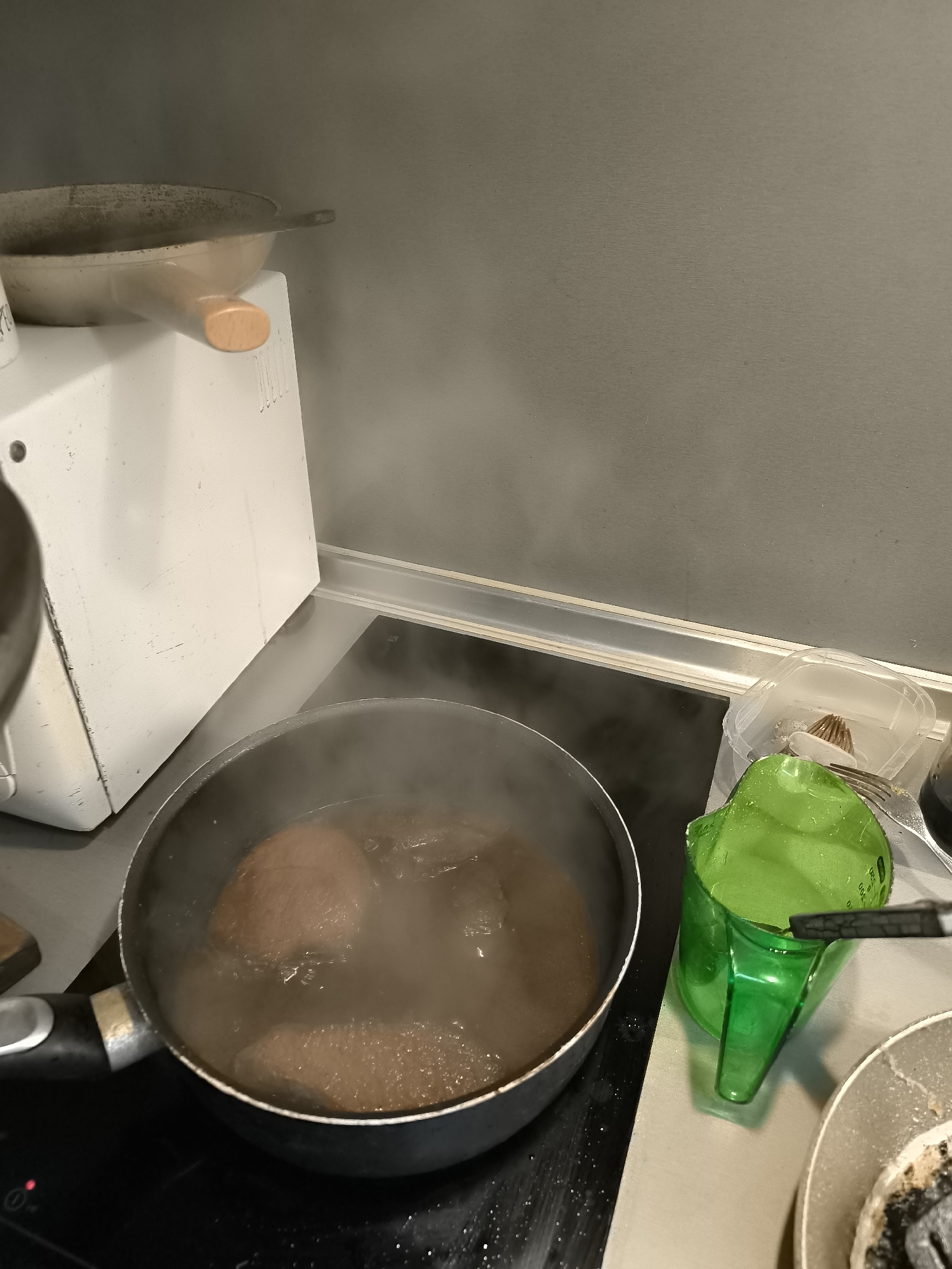 The image shows a kitchen scene. A black frying pan is on a black stovetop, filled with a dark liquid and three round, brown objects resembling meat or large meatballs. Steam rises from the pan. To the left of the pan is a white microwave oven. To the right of the pan is a green measuring cup and a dirty white plate, possibly used for cooking. A plastic container with a fork inside is next to the measuring cup. The wall behind the stovetop is grey. The photograph provides no information regarding the city of Sofia, Bulgaria.