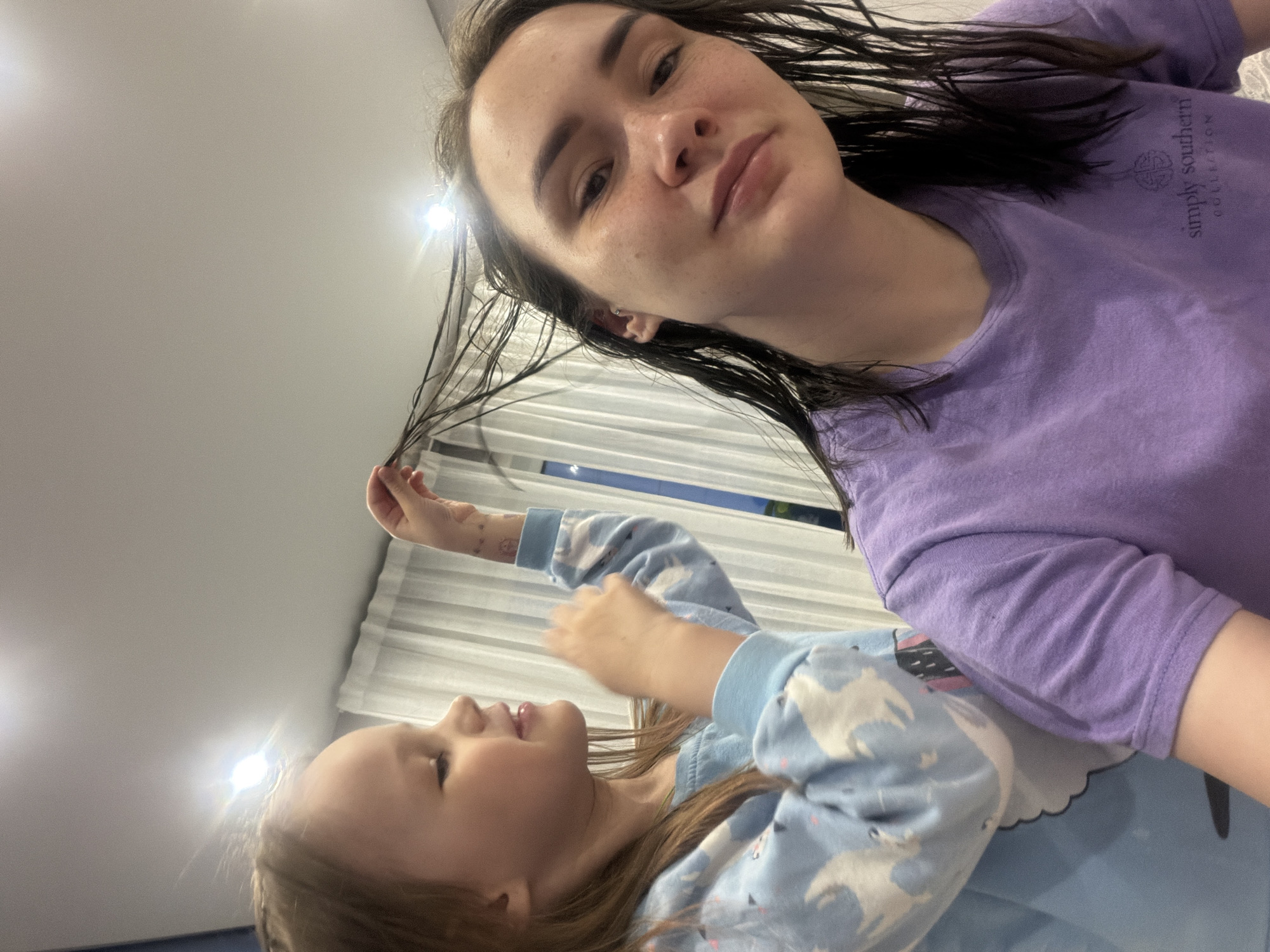 This rotated selfie-style image features an adult female and a young child indoors. The adult, positioned in the upper right, looks directly towards the camera with a neutral expression. She has dark, wet hair, and some freckles are visible on her face. She wears a purple short-sleeved t-shirt with "Simply Southern Collection" text and a logo visible on the right side of her chest. Her left hand is near her hair, appearing to hold a strand.
The child is in the lower left of the frame, looking up towards the adult with a joyful expression. She has light brown hair and is dressed in a light blue long-sleeved top adorned with a pattern of white animals (possibly unicorns or pegasi). Her right hand is raised, and what appear to be temporary tattoos are visible on her forearm. Her left hand rests on the adult's purple sleeve.
The background primarily consists of a plain light-colored ceiling with several bright, glinting light fixtures, suggesting artificial indoor lighting. Behind the subjects, white horizontal window blinds or curtains are visible. The overall scene suggests a casual, intimate moment, possibly at home in Chaikovsky, Russia, as indicated by the location context.
