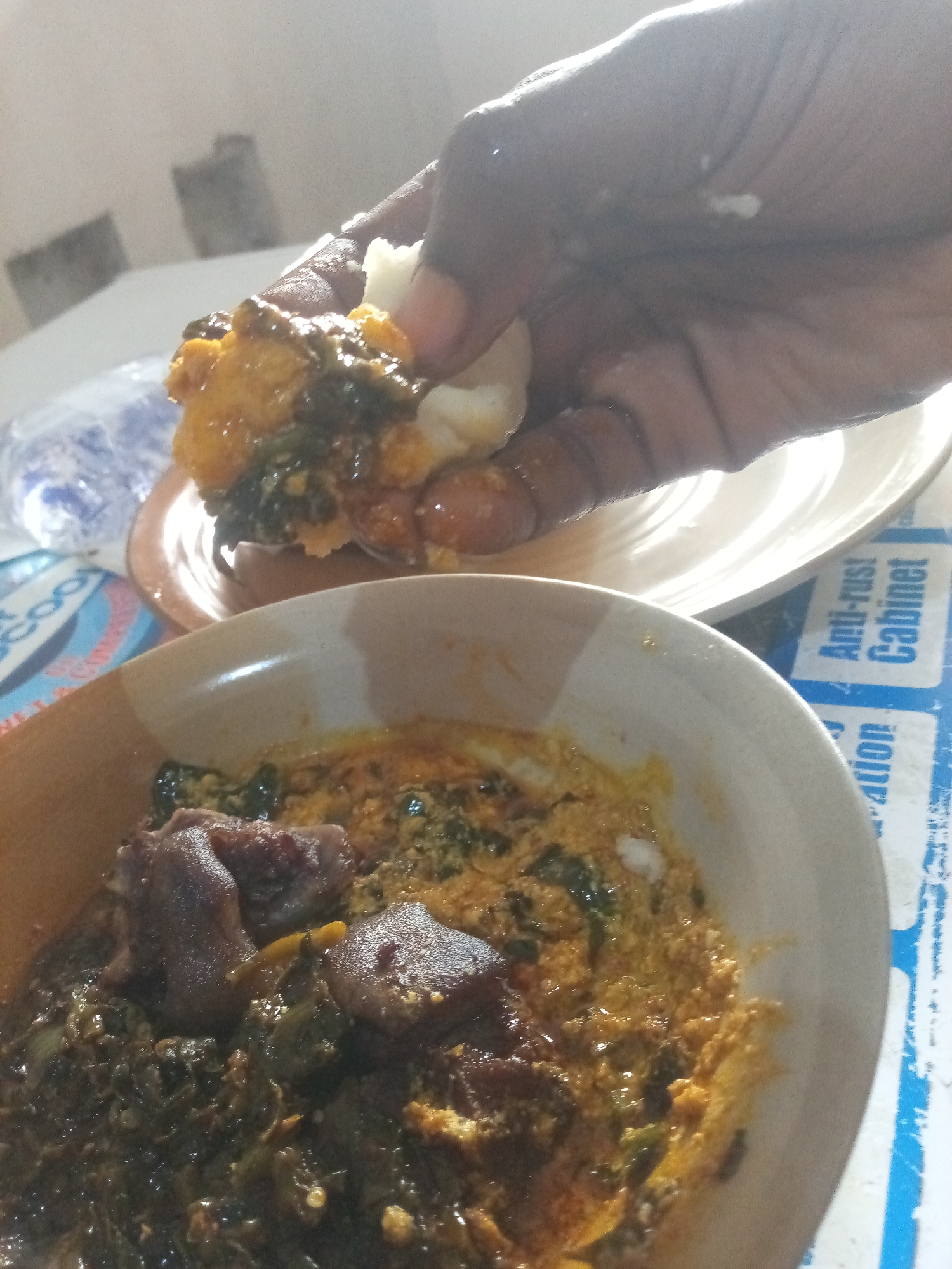 A hand, dark-skinned, is shown picking up a piece of fufu and stew from a plate. The fufu is white and soft, and the stew is a rich, brown color with visible chunks of meat and green vegetables. In the foreground, a bowl of the same stew is visible, with more meat and vegetables. The background is blurred but appears to be a table with a patterned placemat. The setting is likely a home or restaurant, and the food is a traditional West African dish.
