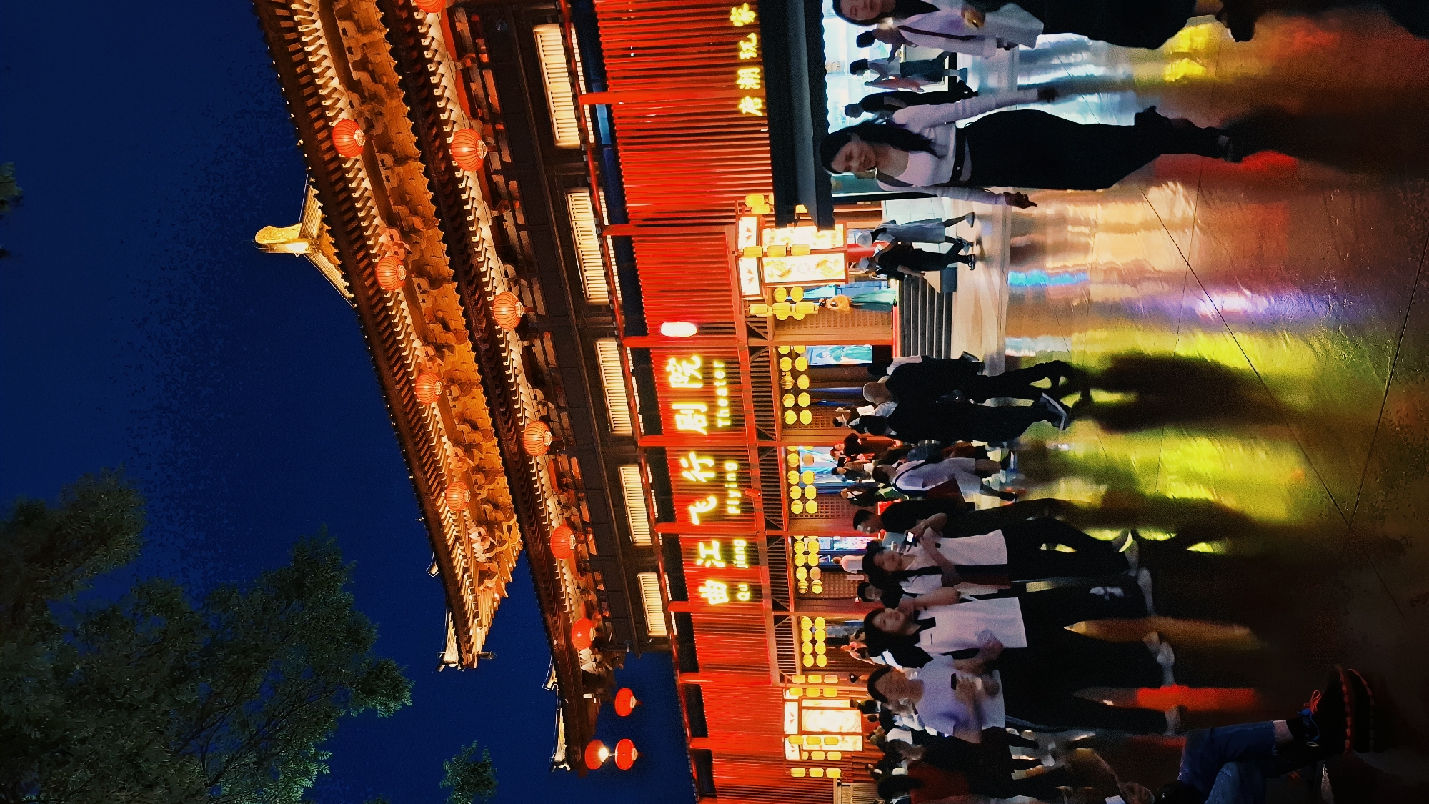 The image shows a crowded area in Yanta District, China, at night. A traditional Chinese building, illuminated with red lanterns, stands on the left. The building's facade is red with lit Chinese characters. People are walking and standing in front of the building, their reflections visible on the wet, brightly lit ground. Some trees are visible on the left side, silhouetted against the dark blue sky.