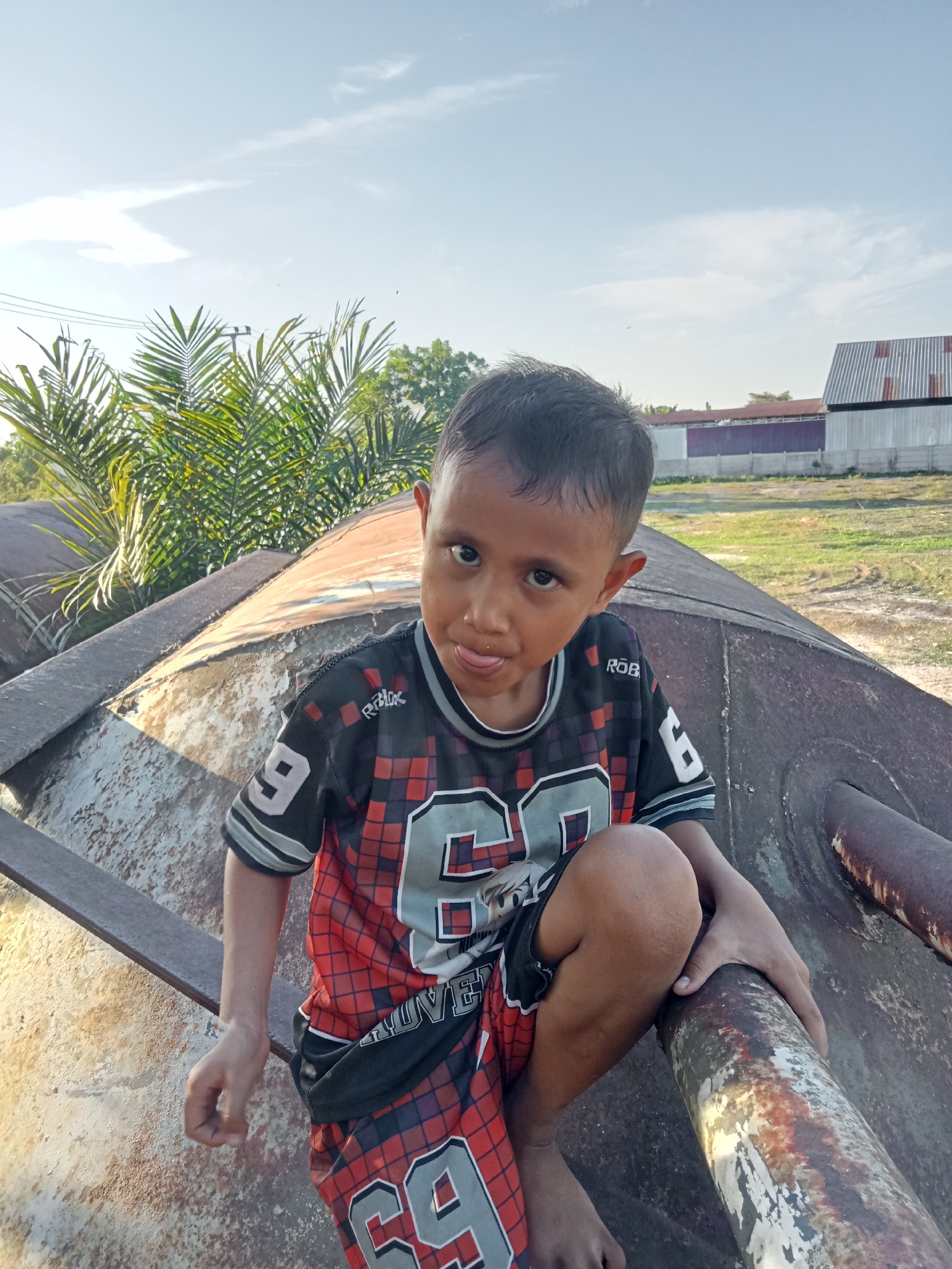 A young boy, appearing to be of Southeast Asian descent, is seated on a large, rusty, cylindrical object in an outdoor setting. He is wearing a black, red, and gray checkered jersey with the number '69' prominently displayed on the front and sleeve, and matching shorts. The boy is looking towards the camera with his tongue sticking out slightly. His right hand is resting on the cylindrical object, and his left hand is near his knee. Behind him, palm fronds and trees are visible, as well as a few buildings with corrugated metal roofs in the distance. The sky above is clear with some wispy clouds, illuminated by sunlight. The location is identified as Kualu, Indonesia. The cylindrical object appears to be made of weathered metal, with signs of rust and peeling paint. The ground in the background is covered with dry grass and dirt.