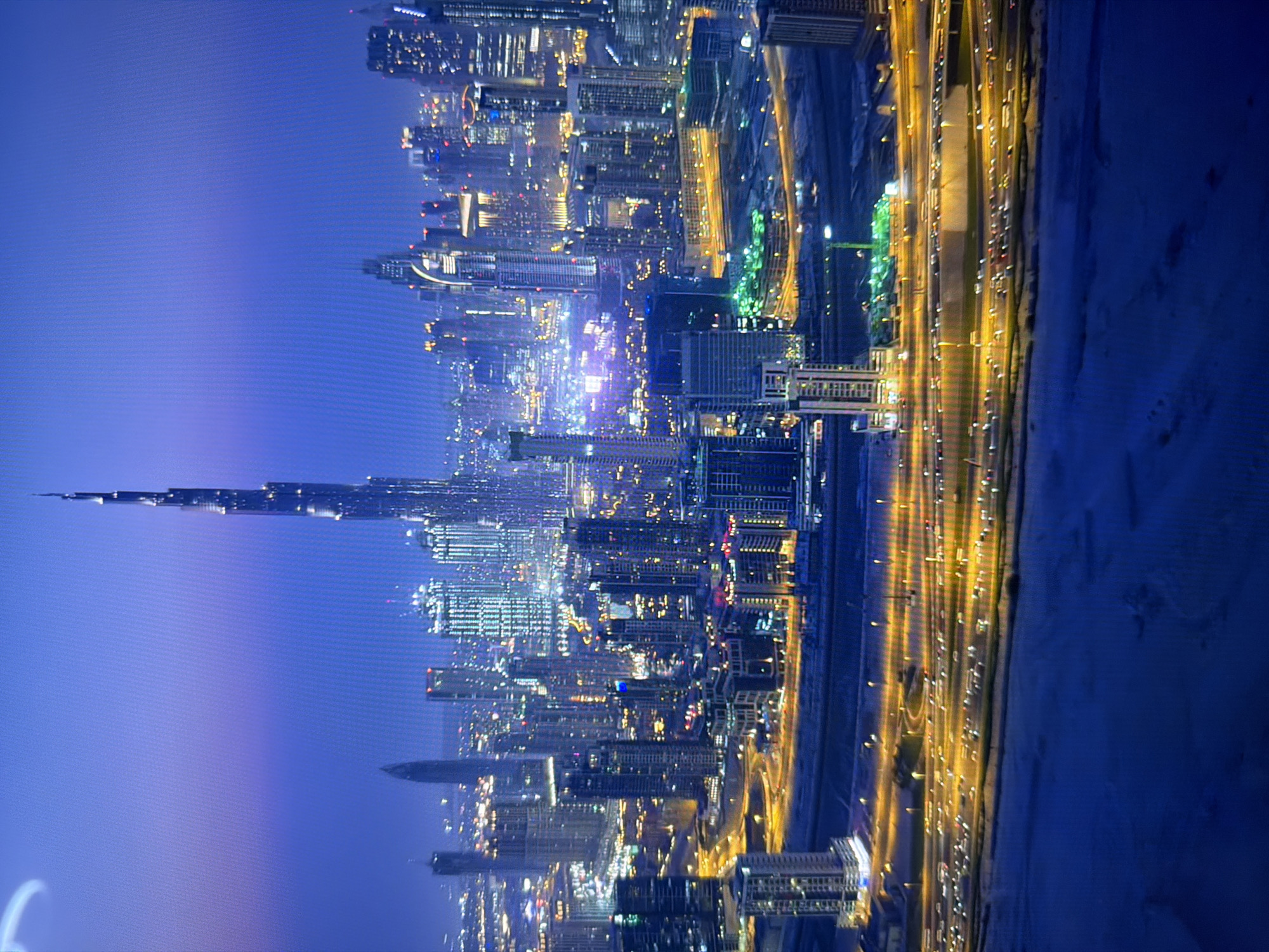 The image shows a nighttime aerial view of a modern cityscape. The most prominent structure is a very tall, slender skyscraper, likely the Burj Khalifa, situated in Dubai, United Arab Emirates. The skyscraper is illuminated and dominates the skyline, surrounded by numerous other high-rise buildings, also lit with various colored lights. A complex network of highways with illuminated vehicles is visible in the foreground, suggesting significant urban development and traffic. The overall scene is characterized by a high density of modern architecture and extensive artificial lighting against a dark, twilight sky. The city depicted is Dubai, not Kyiv, Ukraine.