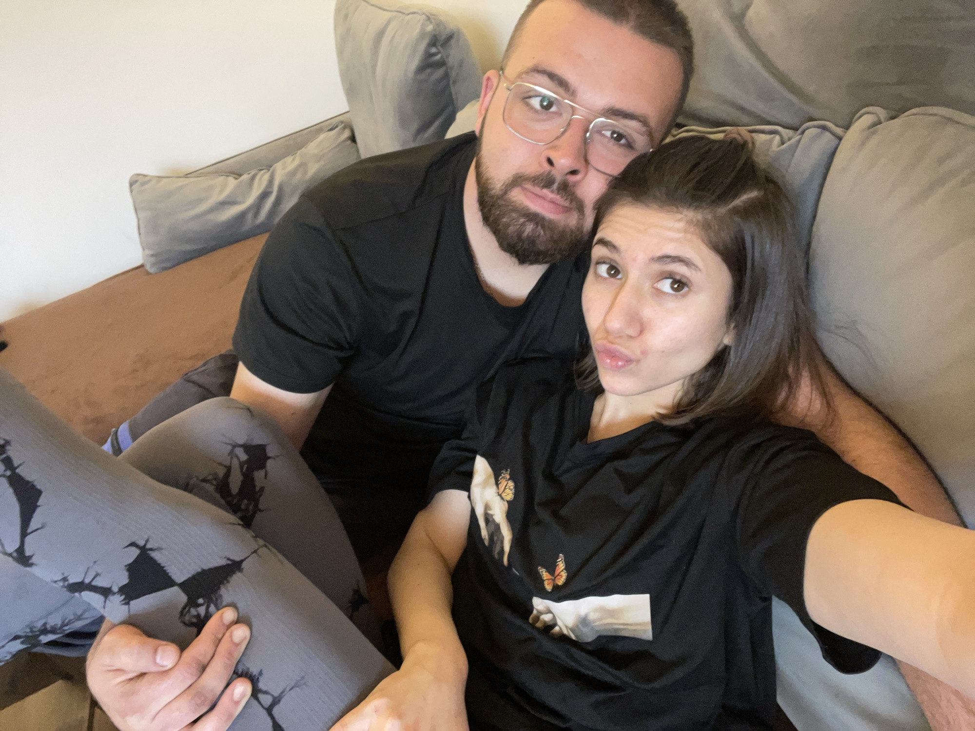 A selfie shows a couple on a sofa. The man is on the left, wearing glasses and a black t-shirt. The woman, on the right, is wearing a black t-shirt with a graphic and grey leggings with a pattern. They are on a brown and grey sofa with several pillows. The background shows a plain wall. There is no indication of the image being taken in Sofia, Bulgaria.
