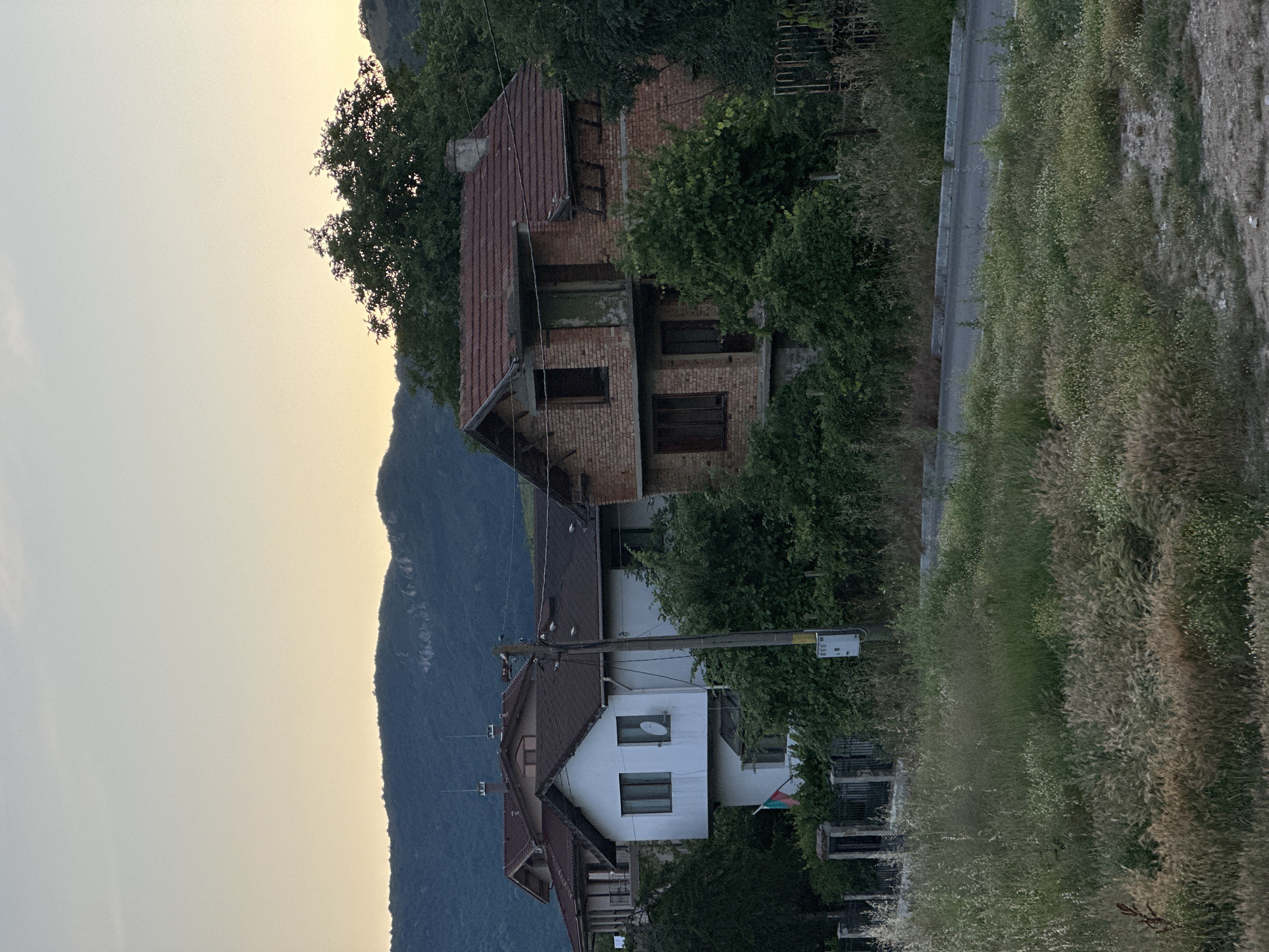 In the city of Unknown, Unknown, a serene rural landscape is captured at dusk. The foreground features a grassy, untamed field with a mix of green and golden hues, suggesting dry vegetation. A paved road curves through the scene, bordered by more wild grass and foliage.
Dominating the midground are several houses. The most prominent is a two-story brick structure, partially in disrepair with visible exposed brickwork and boarded-up windows, indicating it may be abandoned or undergoing renovation. To its left, a white, two-story house with a dark tiled roof stands in better condition, featuring several windows and what appears to be a satellite dish on the facade. A Bulgarian flag is visible near the base of this house. Behind these structures, larger, more modern houses with multiple levels and red-tiled roofs are partially visible.
The background is defined by a dense line of lush green trees that obscure the lower portions of a distant mountain range. The sky above is a soft gradient of pale yellow and light blue, typical of a sunset. The overall atmosphere is peaceful, with a sense of stillness pervading the scene. No people are present in the image.