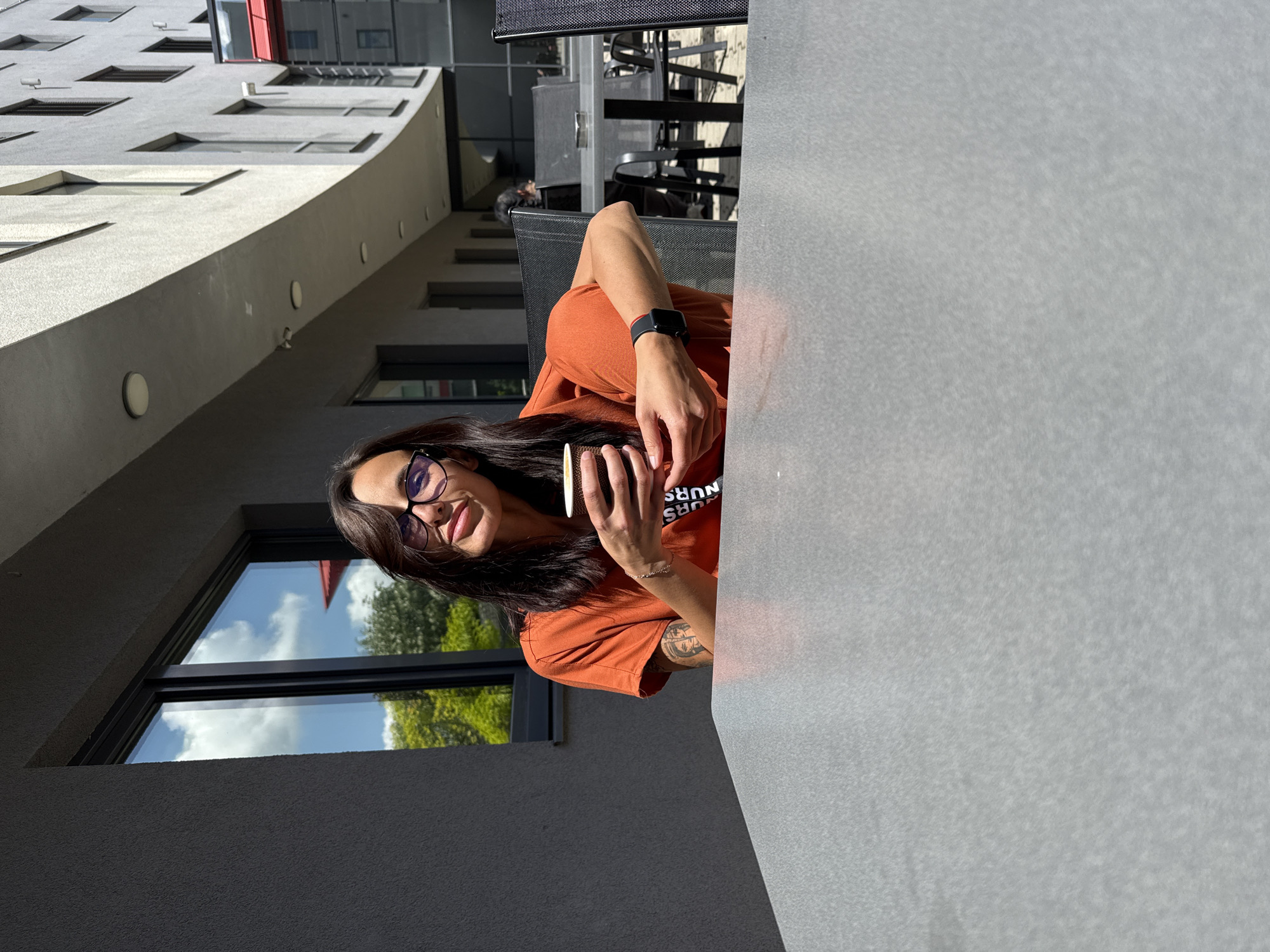The photograph appears to be taken outdoors, likely at a cafe or patio in Pleven, Bulgaria, given the building style and general ambiance. A woman is sitting at a light-grey table, partially visible. She is wearing an orange t-shirt, glasses, and a smartwatch. She is holding a cup and appears to be looking toward the camera. The background features modern architecture with windows reflecting the sky and trees. Several tables and chairs are visible in the background. The light suggests it's a sunny day.