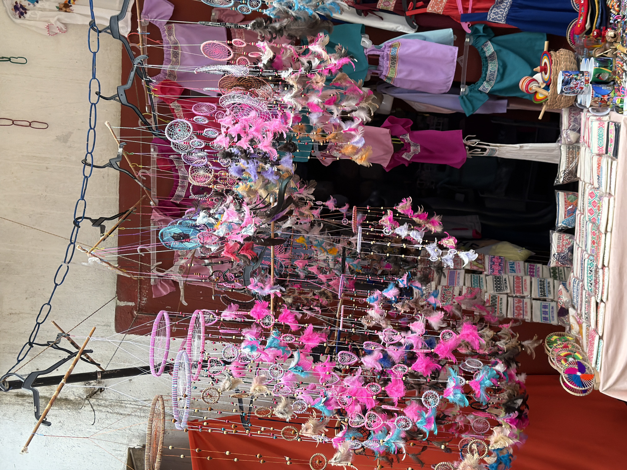 The image shows a market stall in Cuetzalan, Mexico, displaying a variety of crafts. Rows of colorful dreamcatchers with pink, blue, and brown feathers hang on strings, alongside racks of vibrant clothing, including dresses in shades of purple, pink, and teal. Woven baskets containing wooden toys sit next to stacks of embroidered textiles on a pale pink surface, indicating the stall's merchandise for sale. The background includes a faded wall with brick details, chains, and hanging rods, suggesting the stall's setup in a public marketplace.