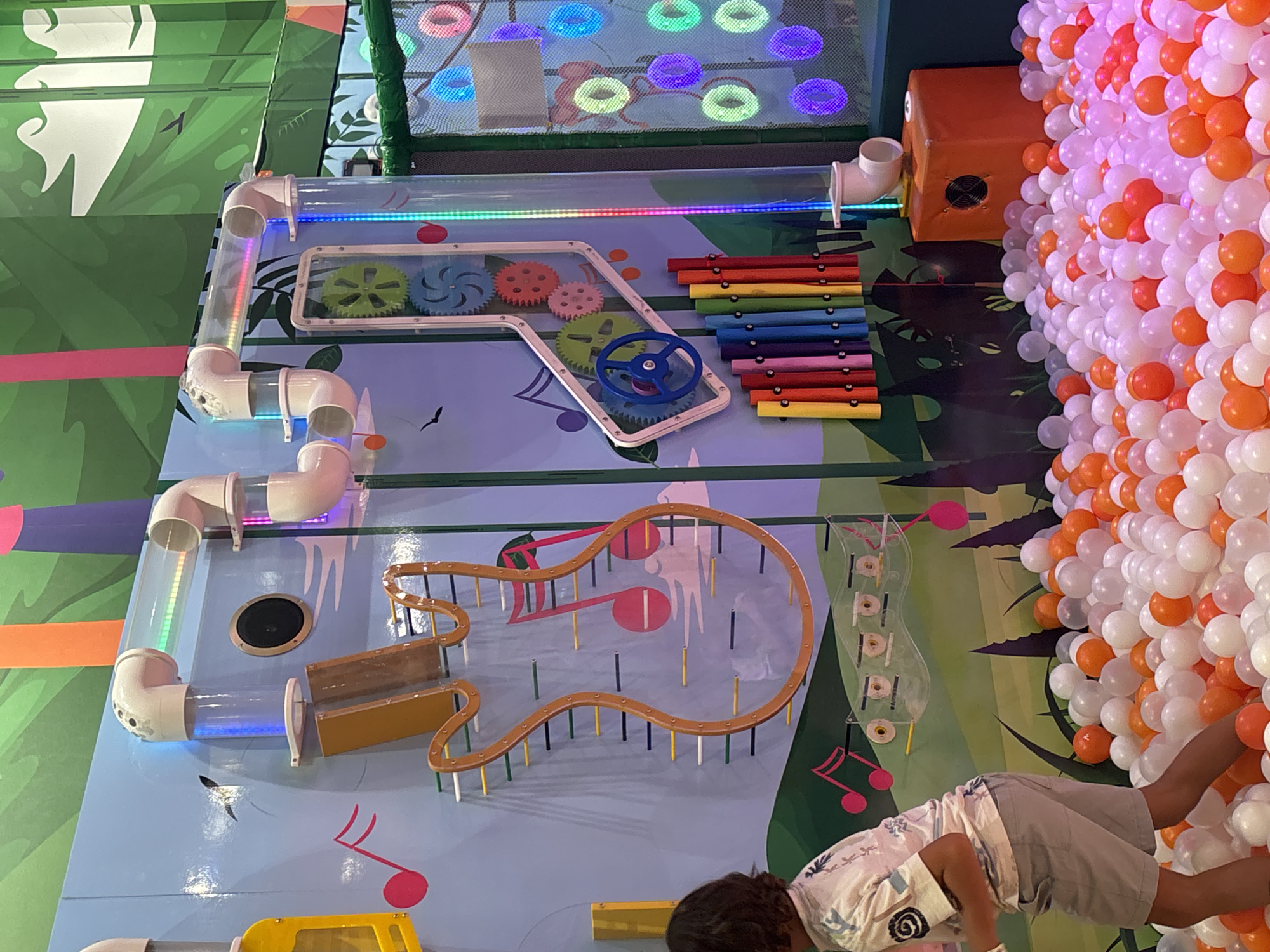 The image depicts an indoor children's play area, likely a ball pit with various interactive installations, set in Dubai, United Arab Emirates. Dominating the scene is a large blue wall adorned with colorful, playful graphics and interactive musical elements. On the left, a young child, partially visible from the back, is wading into a ball pit filled with white and orange balls. They are wearing a patterned short-sleeved shirt and grey shorts.
The interactive wall features a series of clear tubes with white connectors, some containing colorful lights. Below these tubes, a large, clear guitar-shaped frame is mounted, with multiple colored pegs running horizontally across it, suggesting a musical instrument component. To the right of this, a series of colorful wooden xylophone bars are arranged vertically. Further to the right, a tall clear tube with flashing LED lights descends into the ball pit. In the background, there are rotating gears behind a clear panel, and a netted area with illuminated circular lights in various colors (pink, blue, green, purple, yellow). An orange cubic structure with a black ventilation grate is positioned near the ball pit. The overall environment is bright and engaging, designed for children's entertainment.