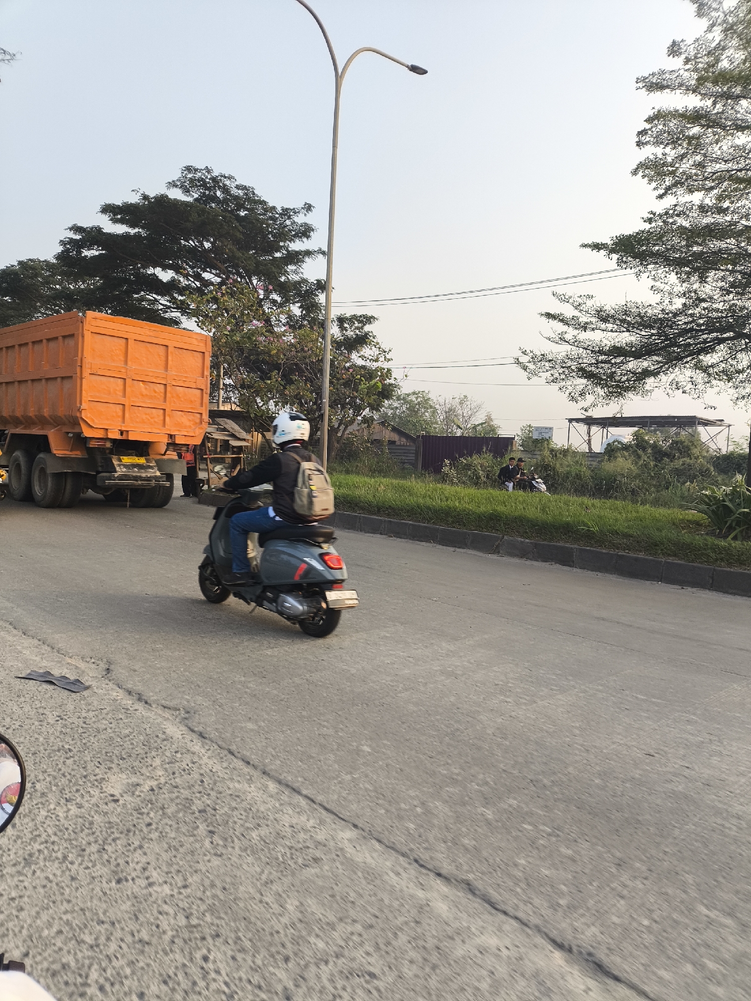 The image depicts a street scene in Cilegon, Indonesia, under a clear sky. In the foreground, a person wearing a white helmet, black jacket, and blue jeans rides a dark gray scooter, carrying a light gray backpack. To the left, a large orange dump truck is parked. The road is paved and shows signs of wear, with some cracks and a black debris item visible on the surface. The background features lush green grass, various trees, some with pink flowers, and vegetation. In the distance, a few people can be seen standing near a small structure, and a billboard is visible amongst the greenery. A tall street lamp with a single light fixture arches over the scene. The perspective suggests the photo was taken from another moving vehicle.