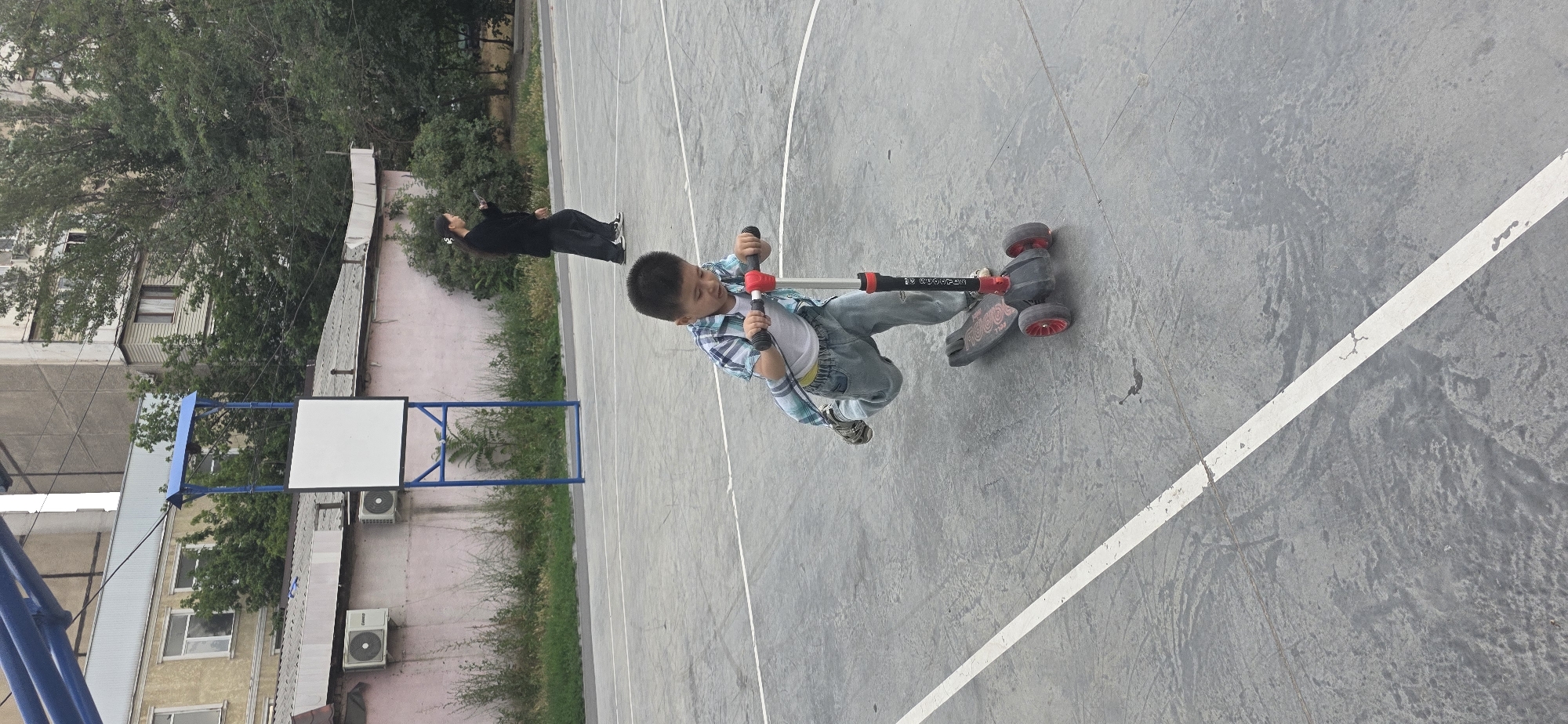 The image shows a young boy riding a three-wheeled scooter on an outdoor basketball court. The boy is wearing a plaid shirt, a white t-shirt, and jeans. He is holding onto the handlebars of the scooter and appears to be in motion. To the left of the boy, another person is visible, lying on the ground and looking up at the sky. The basketball court is made of concrete and has white lines marking the boundaries. In the background, there are buildings and trees. The scene appears to be set in Bishkek City, Kyrgyzstan. The time of day is not explicitly clear, but the lighting suggests it could be daytime. The weather appears to be overcast or cloudy. There is no visible text in the image.