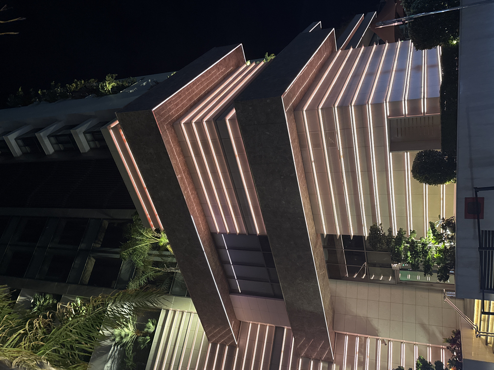 This image shows the exterior of a modern building at night in Agios Tychona Community, Cyprus. The building features a contemporary architectural design with layered facades. The structure is illuminated by horizontal LED strip lights, predominantly in a warm white or light pink hue, which outline the various levels and indentations of the building. These lights create a striking visual effect against the dark night sky.
The building's materials appear to be a combination of dark stone or cladding on the upper, projecting sections, and lighter-colored tiled or concrete panels on the lower, recessed sections. Several large, dark-tinted windows are visible, reflecting the surrounding darkness.
Greenery, including palm trees and smaller shrubs, is incorporated into the building's design, with some plants visible on terraces or balconies and at the base of the structure. The landscaping is also subtly lit, enhancing the ambiance.
In the foreground, a set of stairs with a metal railing leads up to an entrance area. There are no people visible in the image. The overall impression is one of a sophisticated and well-lit urban environment.