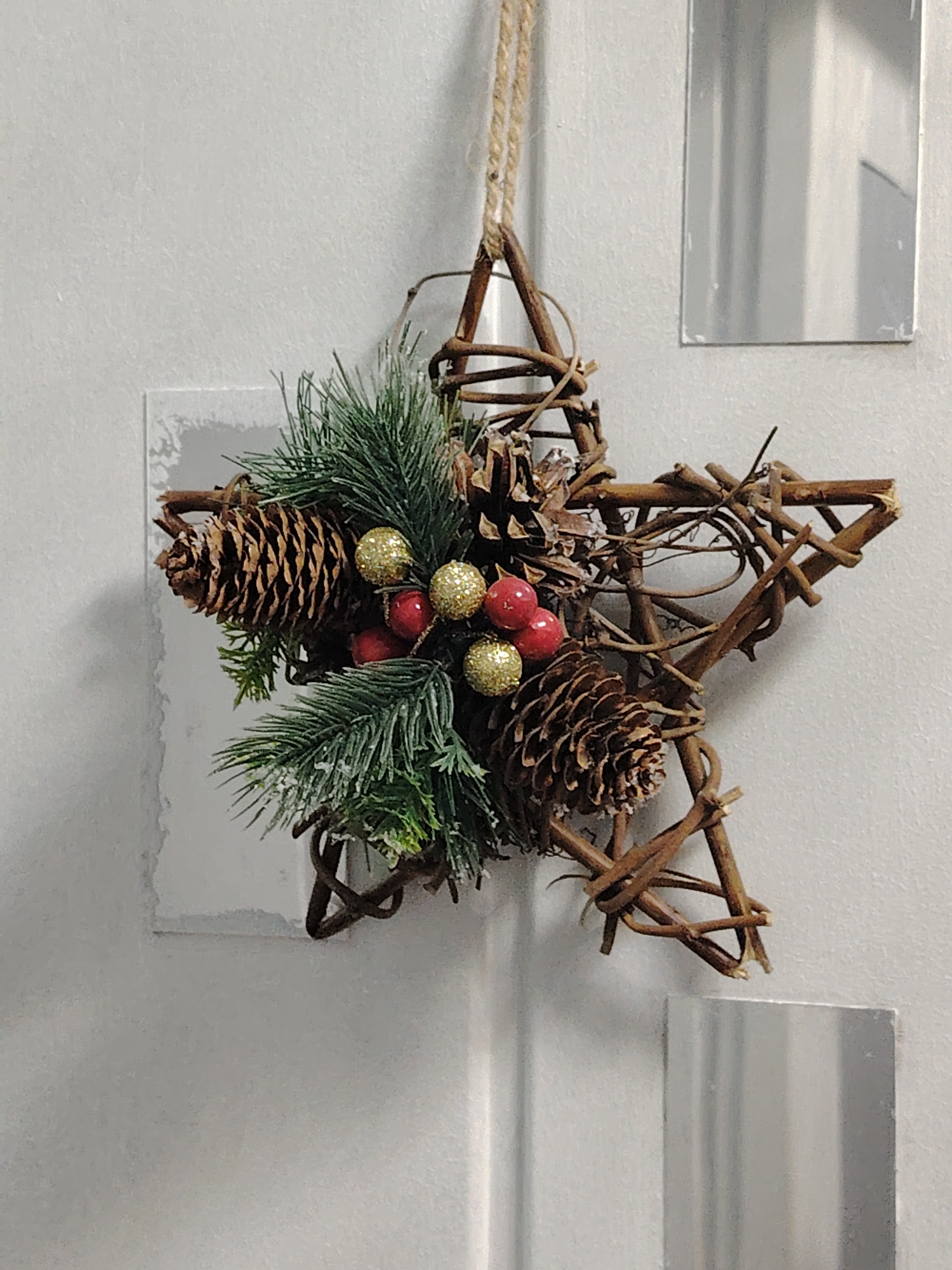 A rustic, twig-formed star ornament, decorated with pinecones, artificial pine sprigs, gold glitter balls, and red berries, hangs from a twine loop against a textured, light-colored wall. Two small, rectangular silver mirrors are affixed to the wall to the right of the ornament, and a larger, square white tile with a grey shadow is visible to the left. The lighting is diffused, suggesting an indoor setting. This scene could be found in a home or shop in Astana, Kazakhstan, as part of a seasonal display.