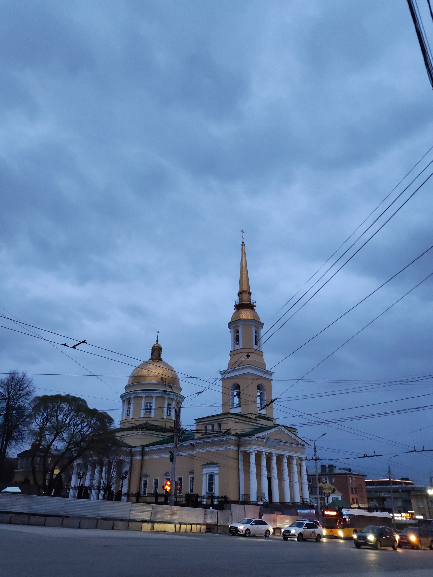 This evening scene in Izhevsk, Russia, features the Alexander Nevsky Cathedral prominently under a dark, heavily overcast sky. The cathedral is a large, light yellowish-beige building with neoclassical architectural elements, including a large golden dome on its left side and a tall, slender golden spire with a bell tower on its right. Columns adorn its facade.
Numerous dark overhead wires crisscross the sky, creating a dense network above the street and in front of the cathedral. In the foreground, an asphalt road shows several vehicles, including multiple cars and a yellow bus, all with their headlights illuminated, indicating the low light conditions of dusk or evening. A red traffic light is visible on the left, near the road.
To the left of the cathedral, mostly bare trees stand, with another building partially visible behind them. To the right, other urban buildings line the street in the distance. A low, light-colored barrier or fence runs along the roadside in the midground. The dense cloud cover contributes to a muted and somber atmosphere. No readable text is visible on any signs, buildings, or vehicles.