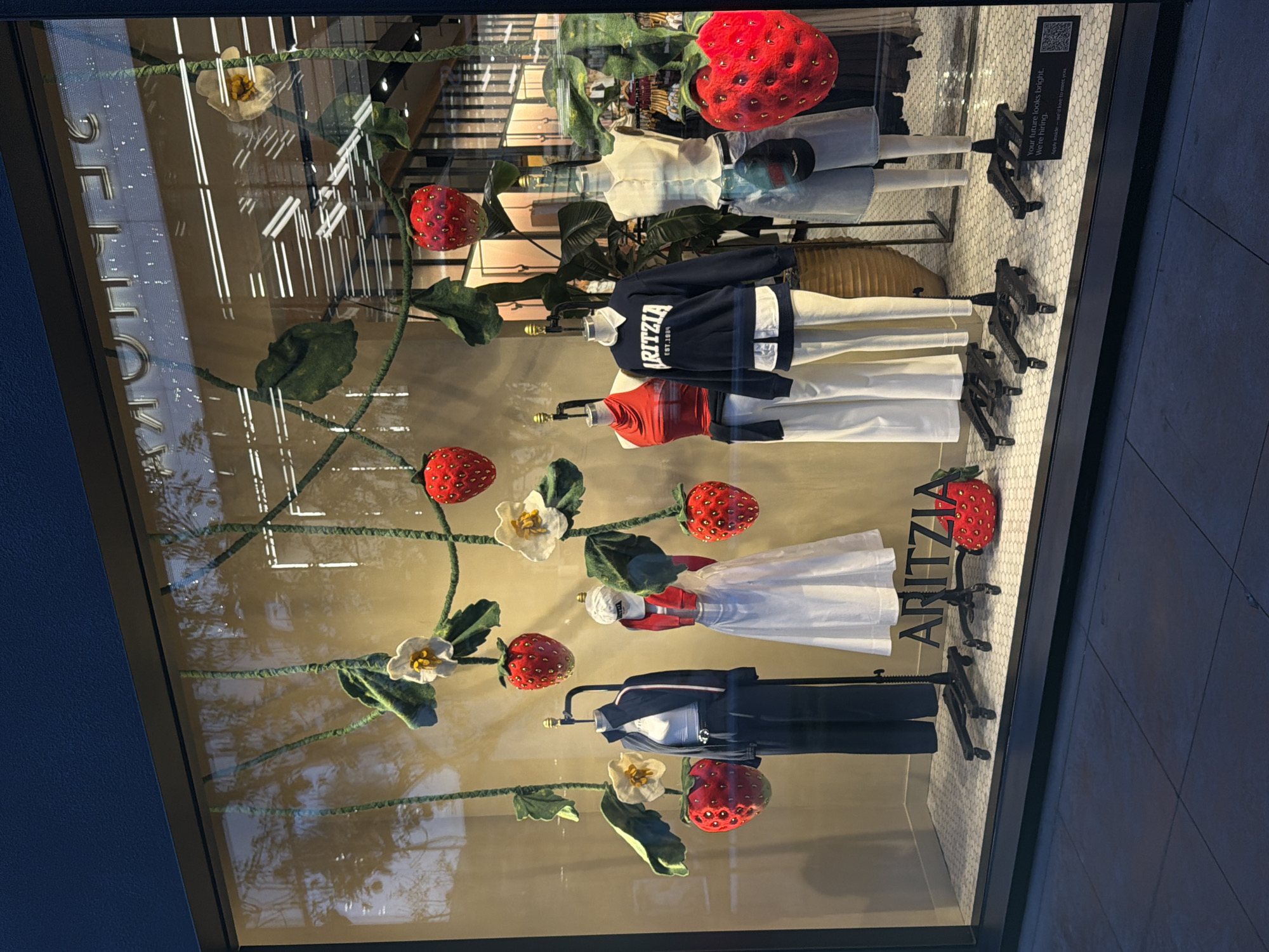 The image shows a retail store window display with a strawberry and floral theme. Several mannequins are dressed in clothing, including pants, tops, and jackets. The display features large, artificial strawberries and white flowers hanging from green vines. The store's name, "ARITZIA," is visible in the display. A sign with a QR code and text reading, "Your future looks bright. We're hiring. Apply inside – we'd love to meet you." is placed in the lower right corner of the window. The display is set against a cream-colored backdrop with horizontal white lines. The window reflects the sky and some trees outside, suggesting it is daytime or early evening. The sidewalk in front of the store is visible, made of grey square tiles. Based on the ARITZIA store branding and the style of the display, this is likely a storefront in a commercial area. Without further context, the exact location in Los Angeles, USA cannot be determined, but the clean, modern aesthetic suggests a location in a shopping district.