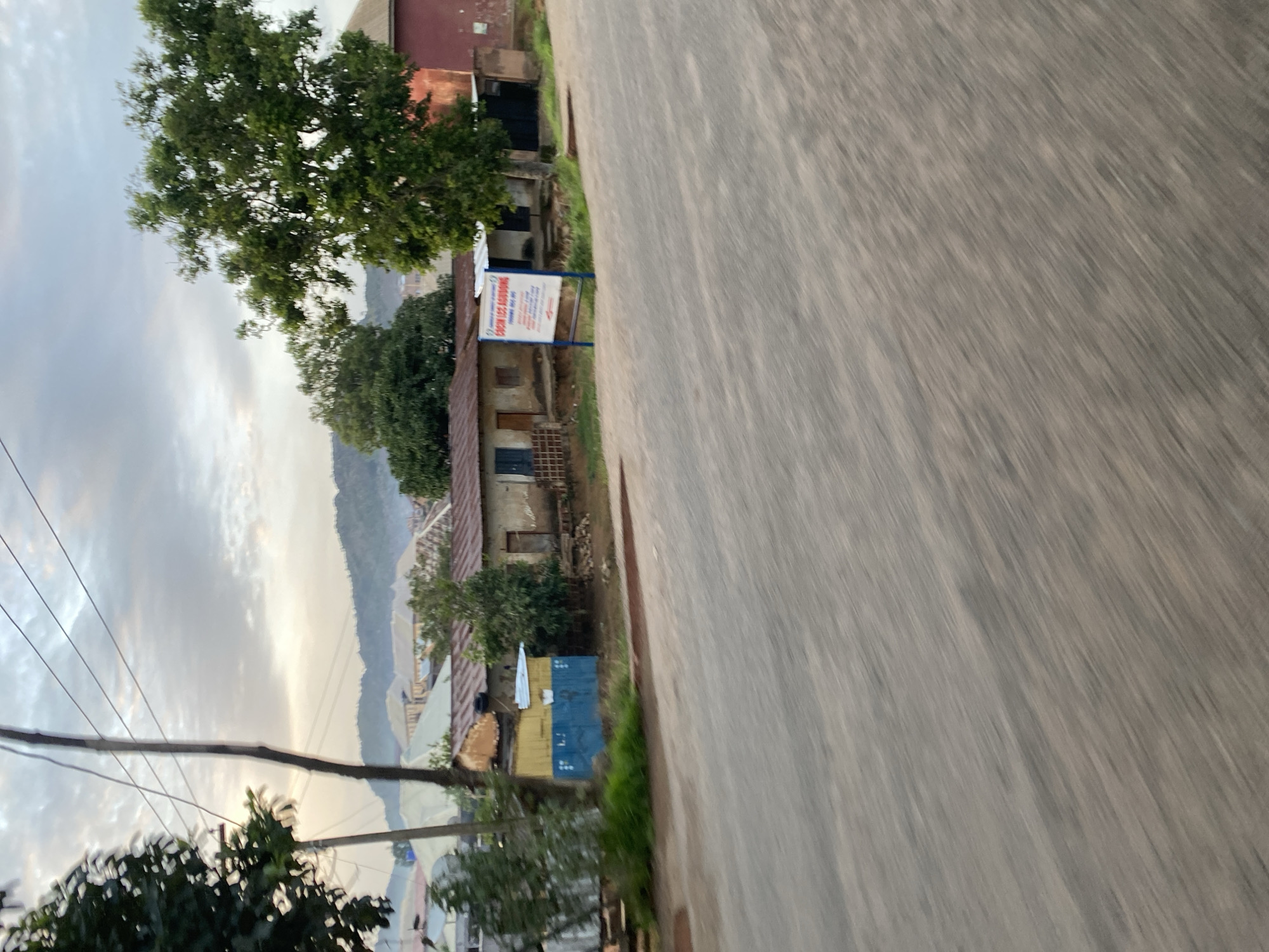 The image depicts a street scene in Jos, Nigeria, during what appears to be early morning or late evening due to the soft, diffused light and cloudy sky. The foreground is dominated by a blurred, grey asphalt road, indicating movement. Behind the road, a low-rise building with a corrugated metal roof is visible. Adjacent to it is a smaller, yellow and blue structure, possibly a kiosk or storage unit. Further back, more buildings with similar roofing materials are seen, set against a backdrop of rolling hills that are characteristic of Jos. A large tree with green foliage stands to the right of the frame, and another tree is partially visible on the left. A white sign with blue and red text is planted in front of one of the buildings. The overall impression is of a typical urban or semi-urban environment in Nigeria, with modest structures and natural vegetation.