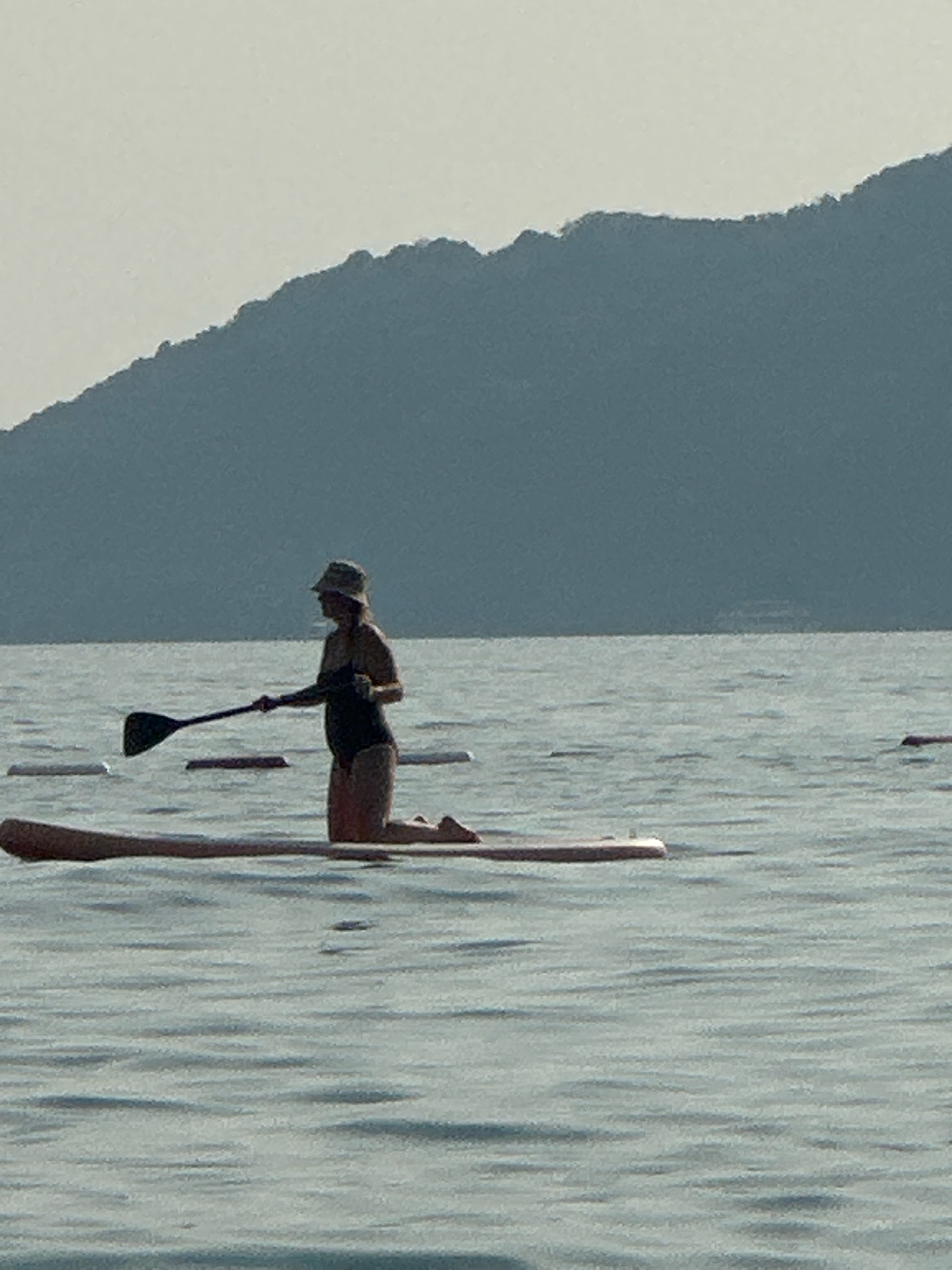 A person is kneeling on a paddleboard in the water, holding a paddle. The person is wearing a swimsuit and a hat. In the background, there is a mountain range. The scene appears to be a serene body of water, likely a lake or calm sea, under a hazy sky. The image depicts a solitary activity in a natural setting. The location is not specified as Boreti, Montenegro, as this information is not present in the image or its provided context.