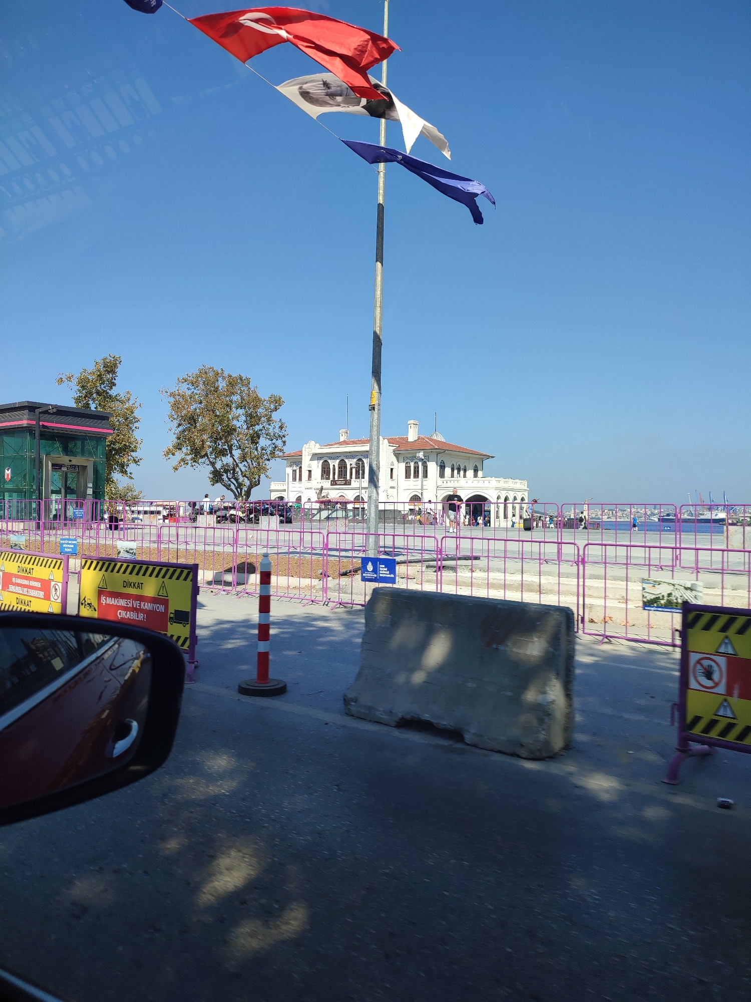 The image shows a bright, sunny day in Kadıköy, Turkey, likely near the ferry terminal. In the foreground, a red car's side mirror reflects the scene. A gray concrete barrier is visible, along with construction fencing and warning signs. A red and white striped pole stands near the barrier.
In the mid-ground, a white building with a distinctive architectural style, likely a historic ferry terminal, is prominent. Flags are strung from a tall pole in front of the building. Trees with green and brown foliage are present. People are visible walking around the building and on the promenade.
In the background, the sea is visible with several ships and cranes. The sky is a clear, vibrant blue. The overall impression is one of a busy, public area with ongoing construction or renovation.