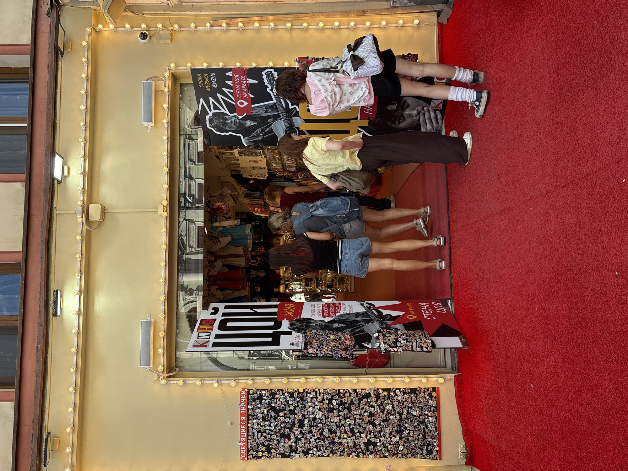 The image shows the entrance to a shop in Moscow, Russia, decorated with a red carpet. The shop is called "Кино Цоя" and features a large poster of Viktor Tsoi, a famous Russian rock musician. The shop sells merchandise related to Tsoi, such as t-shirts, pins, and posters. Several people are standing in front of the shop, looking at the merchandise. The shop is illuminated by string lights, which create a festive atmosphere. The building has a yellow facade and is located in an urban setting.