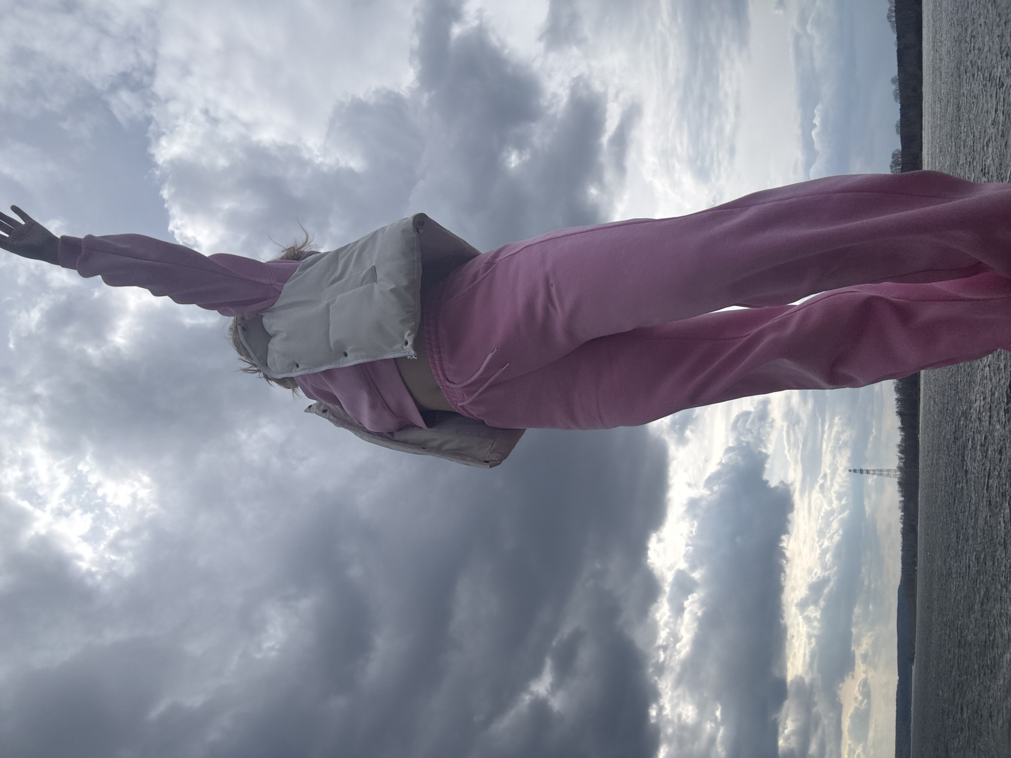 The image, originally oriented 90 degrees clockwise, depicts a person with outstretched arms against a dramatic sky and waterscape. When viewed upright, the subject, likely female, is seen from a low angle, with their arms spread wide in a gesture of openness or freedom. They are dressed in a pink tracksuit and a light-colored (possibly beige or cream) puffer vest. Light-colored hair, appearing somewhat windblown, is visible around their head.
The setting features a vast, overcast sky filled with dark grey and white clouds, with brighter patches indicating sunlight breaking through. To the right of the person, a body of water, likely a river or lake, stretches into the distance, its surface rippled. Beyond the water, a distant shoreline is visible, marked by a line of bare trees. A tall, slender structure, possibly a transmission tower or communication mast, stands on this far bank. The scene suggests daytime, under cloudy weather conditions, possibly conveying a sense of peace or liberation. No visible text is present in the image. The location is Chaikovsky, Russia.