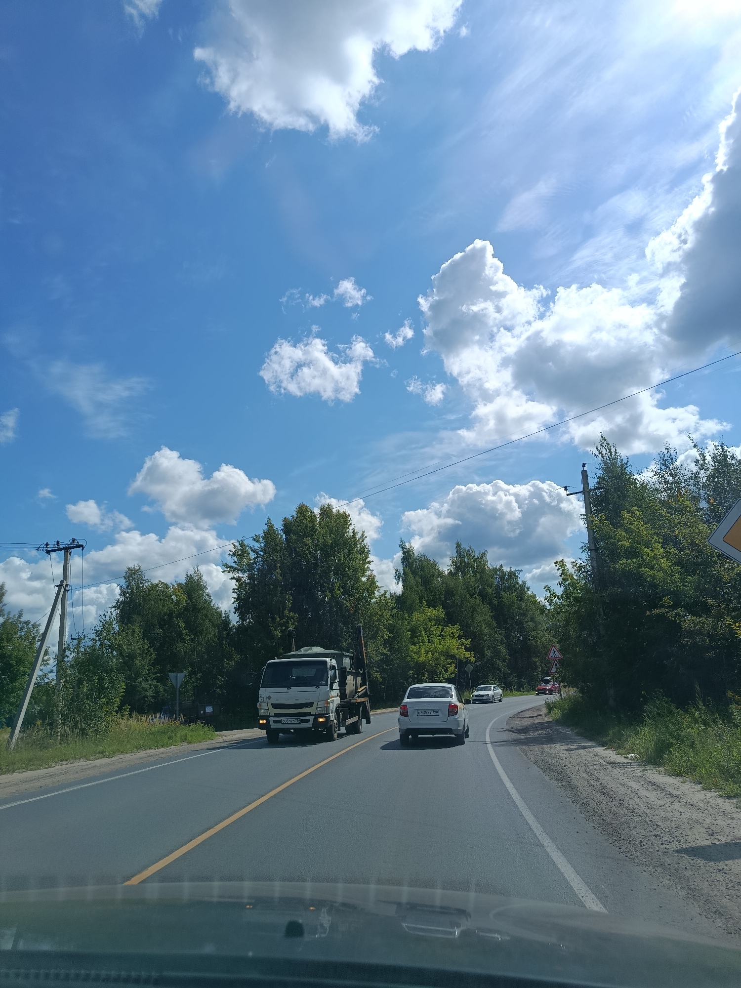 A daytime, eye-level, exterior shot captures a two-lane asphalt road winding through a treed area under a bright blue sky dotted with white cumulus clouds. A white truck with a dark-colored bed, carrying what appears to be sand or dirt, is traveling in the right lane, facing away from the camera. Further ahead, a white sedan is also moving in the right lane, followed by two other vehicles further in the distance. The road is bordered by green grass and dense trees on both sides, with a single utility pole and wires visible on the left. The lower portion of the image shows the dashboard and windshield of a vehicle from which the photograph was taken. The scene is consistent with a rural or suburban setting in Тополиный, Russia.