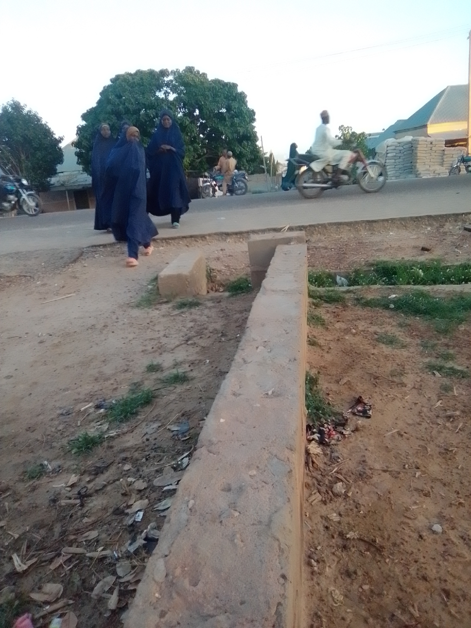In Gombe, Nigeria, four individuals are walking along a dirt road near a paved street. Three of the individuals, appearing to be women, are wearing dark blue hijabs and robes, with one of them also wearing pink sandals. The fourth person is partially obscured. A man wearing a light-colored tunic and trousers is riding a motorcycle in the background, which is a common mode of transportation in Gombe. Also visible are motorcycles parked along the street, buildings with pitched roofs, and stacks of bagged materials, possibly cement, indicating a commercial or construction area. The environment is dusty and arid, with sparse vegetation and debris scattered on the ground, typical of many urban areas in Nigeria.