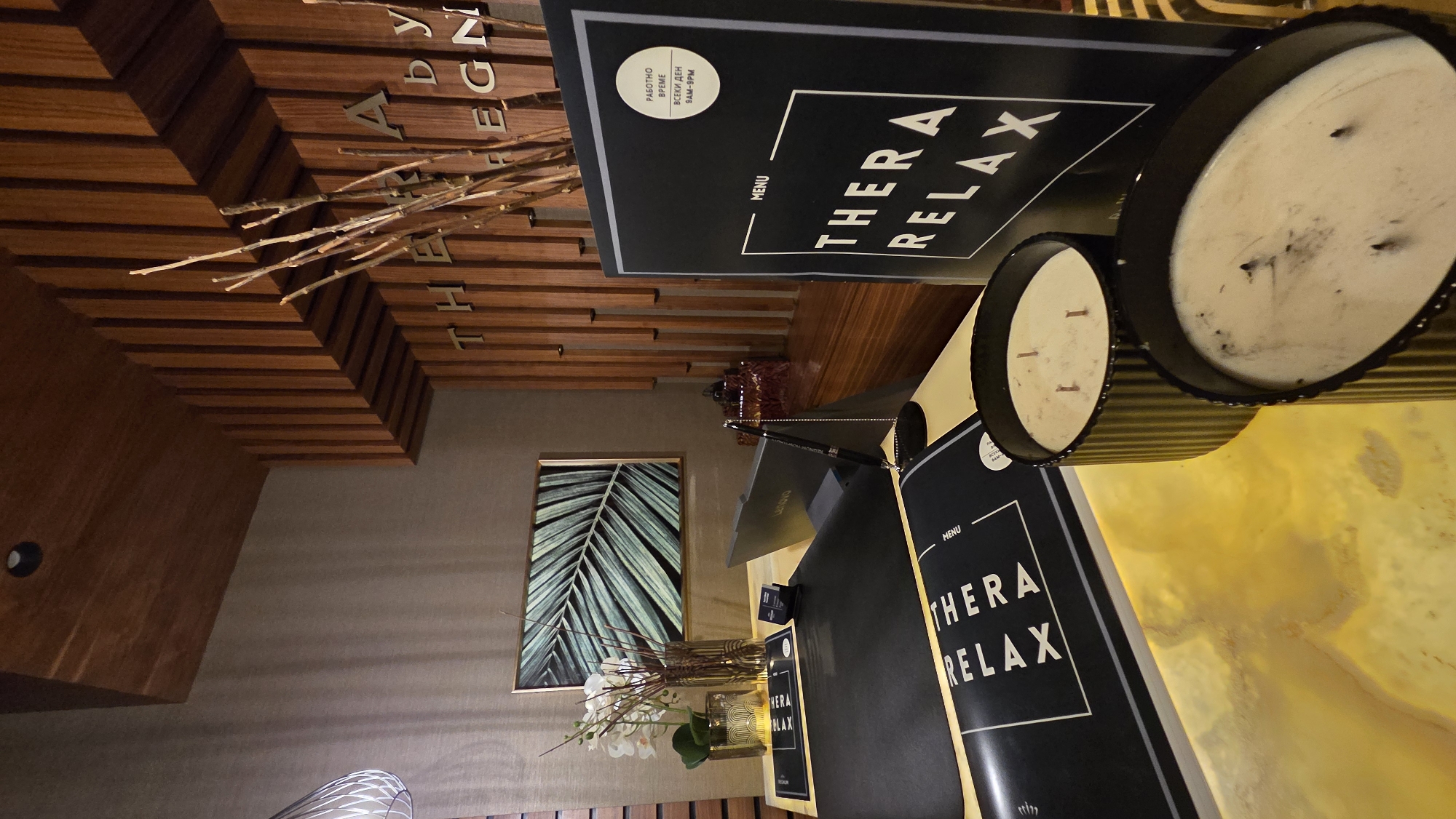 The image depicts a reception or waiting area of a spa or wellness establishment. In the foreground, a counter is set with two large, lit candles in black ribbed containers and a black menu stand with the text "MENU" and "THERA RELAX" prominently displayed. To the left, a black placemat on the counter also displays the text "THERA RELAX" in white lettering. Behind the counter, a laptop computer, a small business card holder, a pen on a stand, and a vase with white flowers and decorative twigs are visible. On the wall behind the counter, a framed artwork of a palm leaf is displayed. The background features a wall with vertical wooden slats. The overall ambiance is modern and serene, with warm lighting and natural elements. The location is not specified.