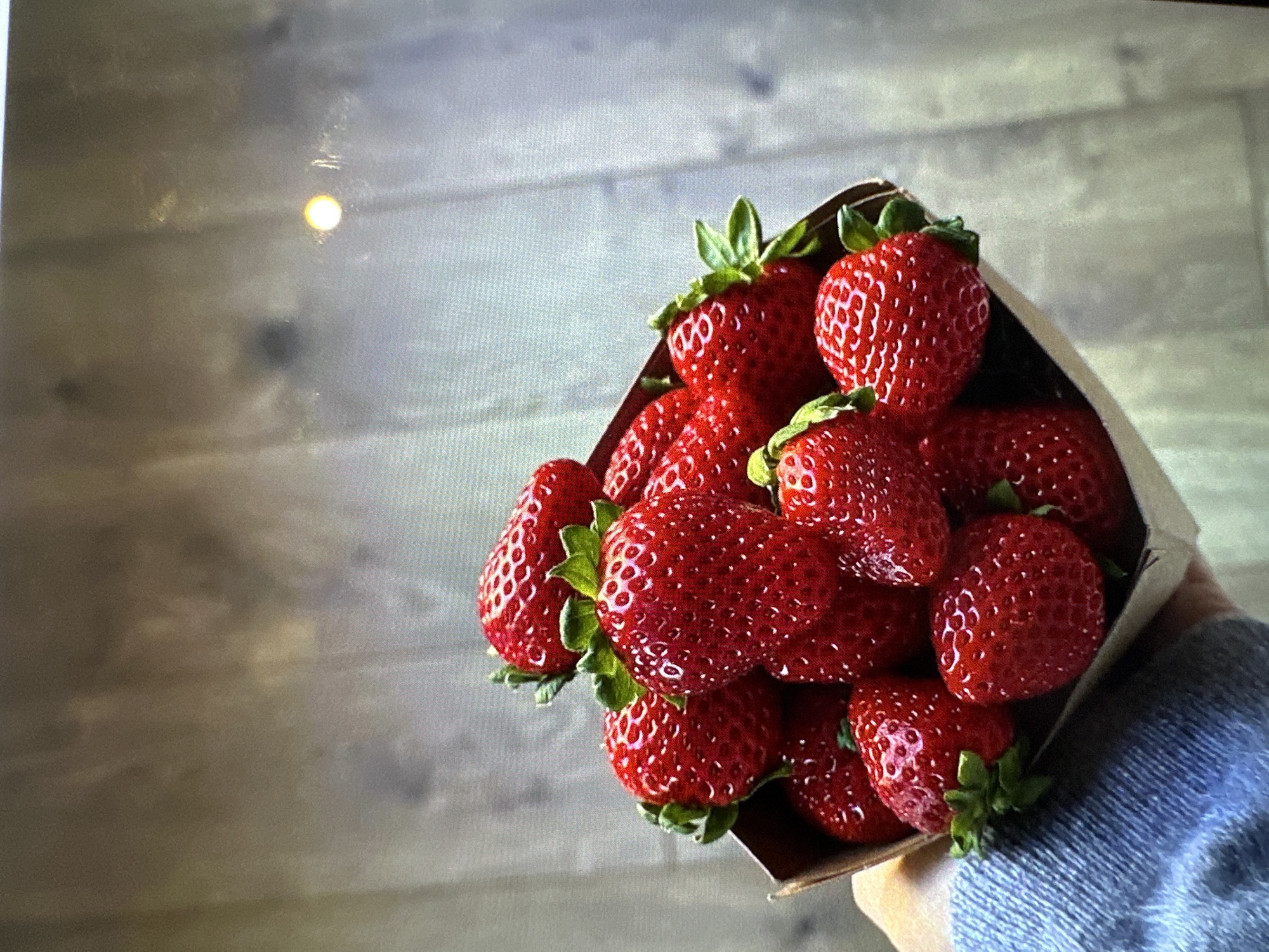 A close-up shot shows a hand holding a brown paper cone filled with fresh, red strawberries with green leafy tops. The strawberries are the main subject, filling most of the frame. The background is a light gray wall with horizontal wood-like patterns. The sleeve of a blue denim shirt is visible on the bottom right. The scene is indoors with sufficient light. The image doesn't provide information to determine if the scene is set in Yerevan, Armenia.