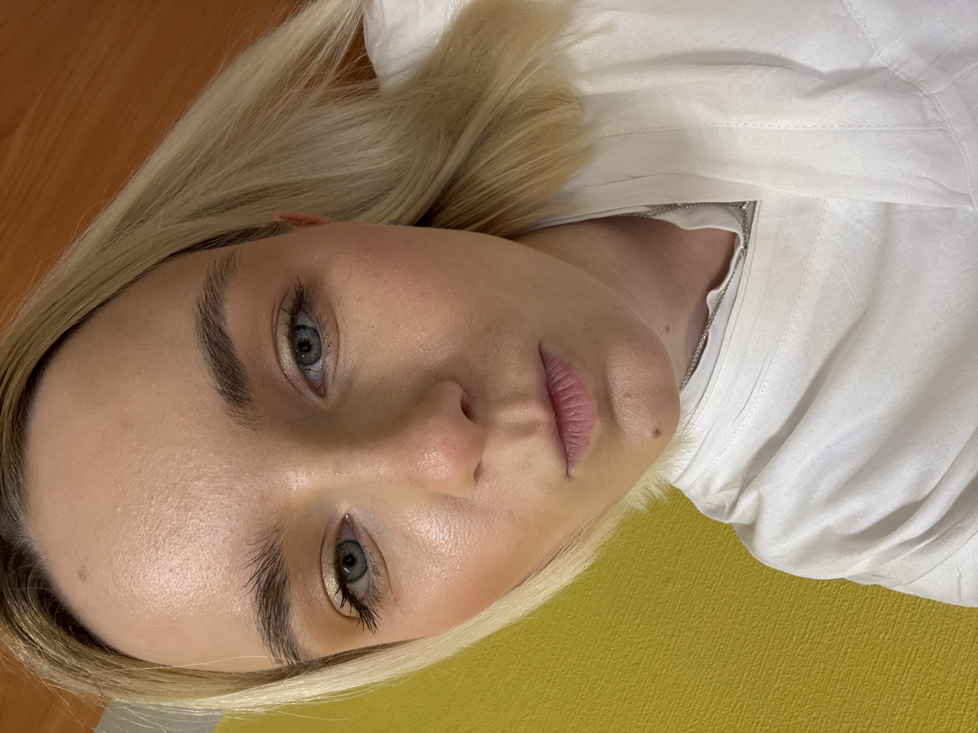 A close-up photograph shows a woman with blonde hair and blue eyes lying on a yellow surface. She is wearing a white collared shirt and a silver necklace is visible. The background includes what appears to be a light brown, possibly wooden, surface. There is no indication in this image that it was taken in Saint Petersburg, Russia.