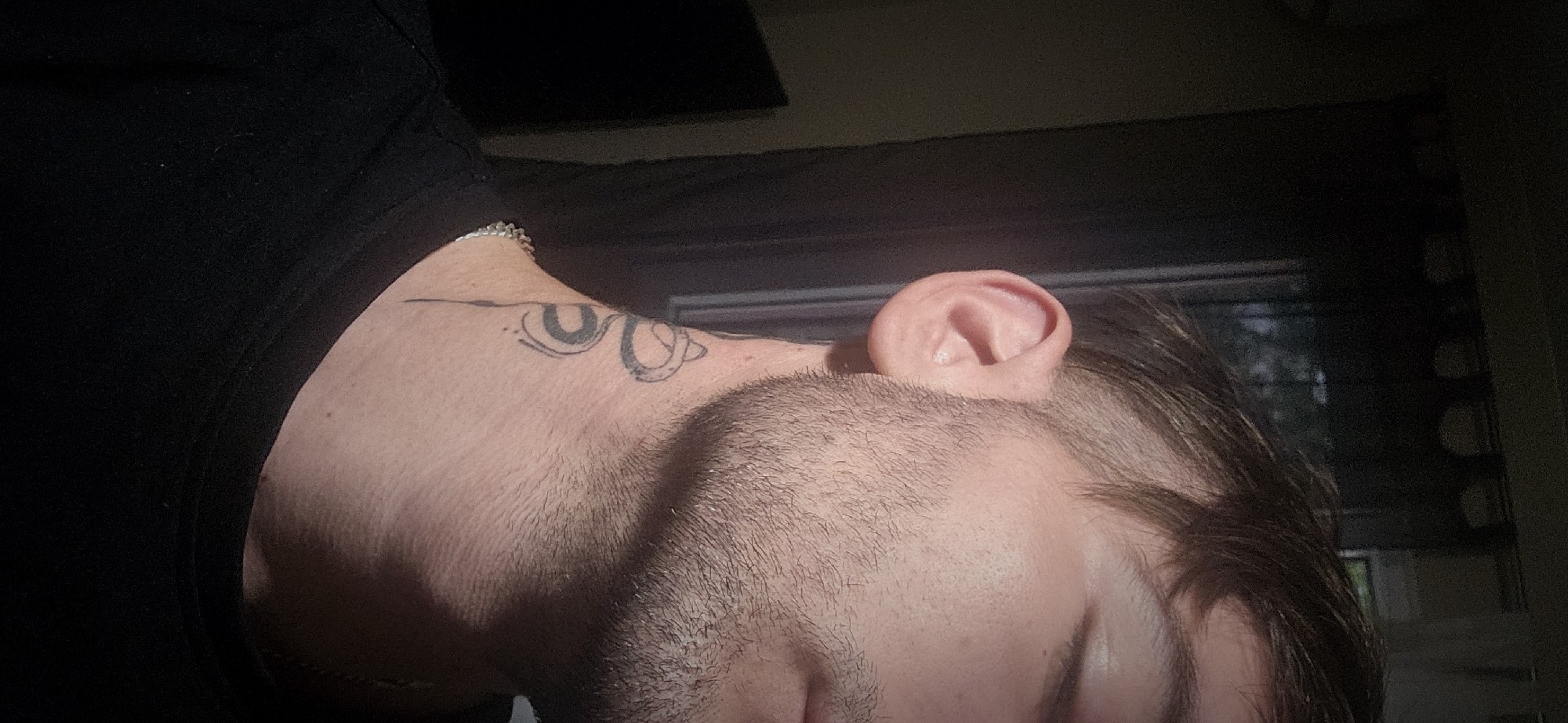 A close-up, low-angle shot features a light-skinned person's head and neck, angled downwards. The person has short, dark hair and stubble. Part of their face, including an eye, is visible. The right ear is prominent. A tattoo is visible on the side of the neck, depicting a stylized design in black ink. The person wears a black t-shirt. A thin chain is visible around the base of the neck. The background is blurred, showing a window and what appears to be either a closed shade or a dark wall. There is no specific indication or element in the image suggesting the location is Plovdiv, Bulgaria.