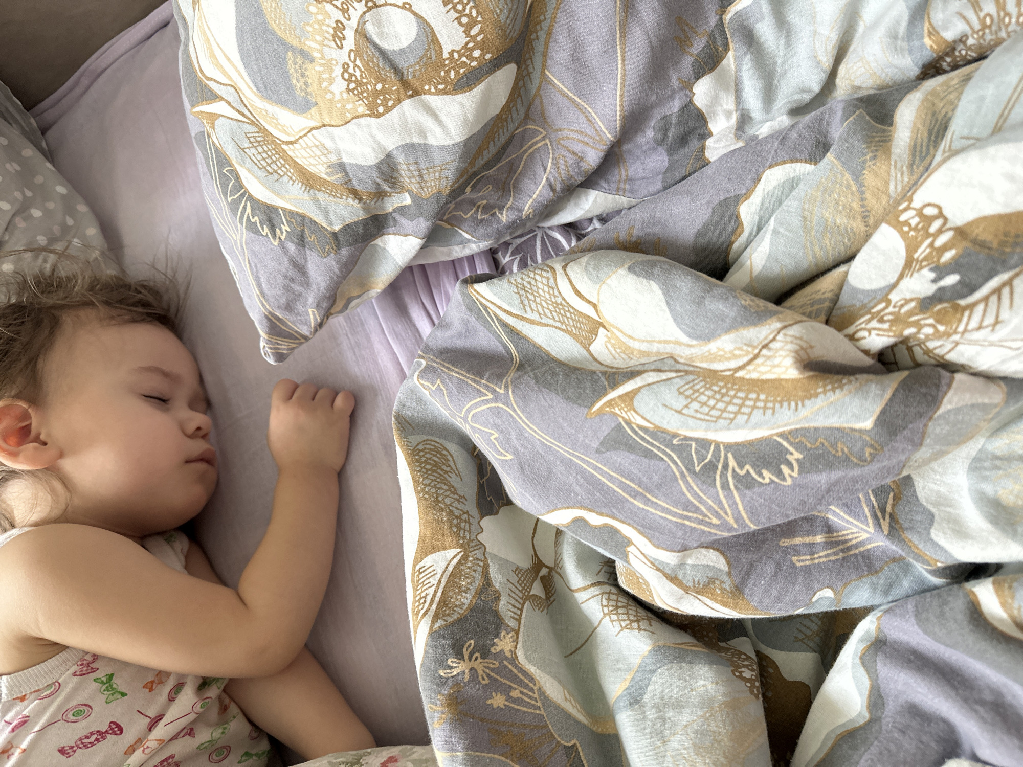 A young child sleeps on a bed covered in lavender linens with a floral-patterned duvet. The child lies on their left side, their right arm raised slightly, with the hand near their head. The duvet is rumpled, revealing the lavender sheet underneath. The scene appears to be indoors, but there is no indication of its location in Uva, Russia.