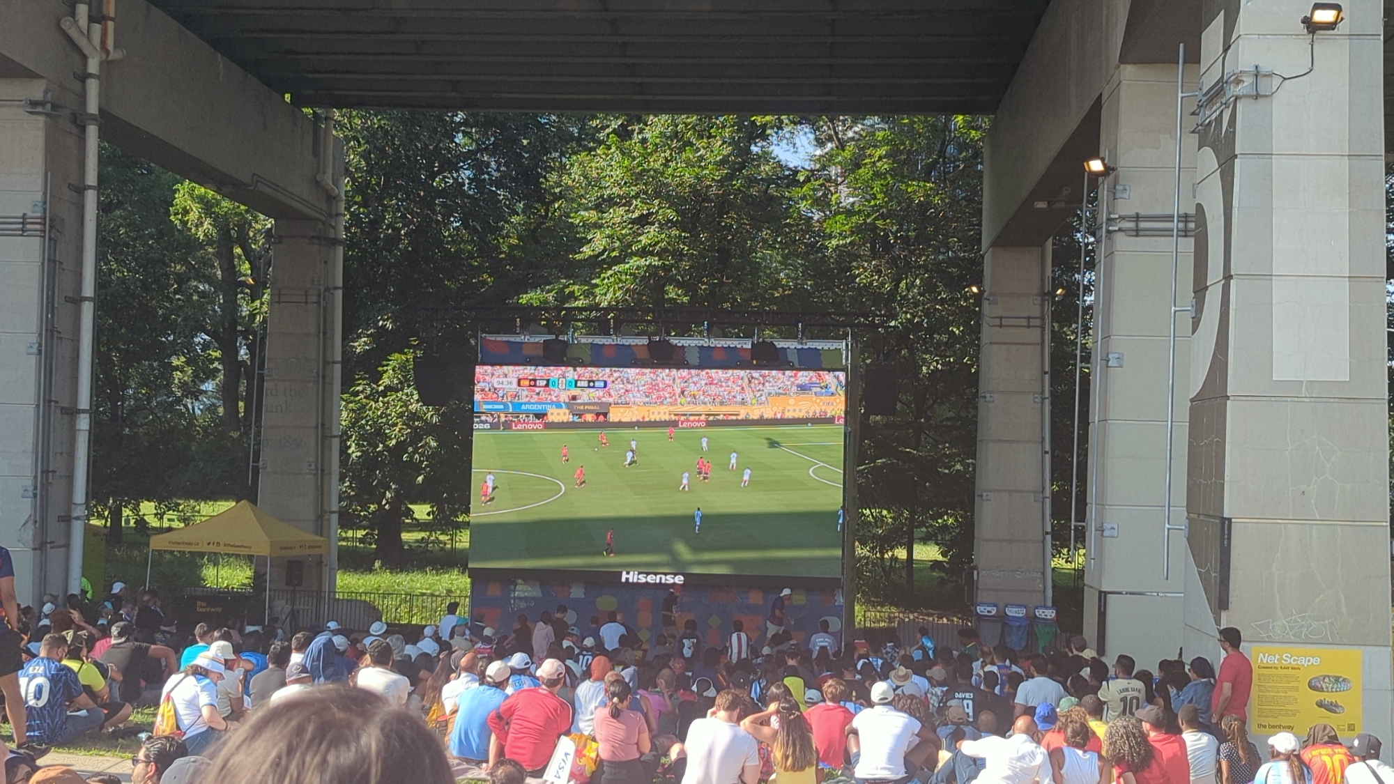 The image depicts a crowd of people gathered outdoors, watching a large screen displaying a soccer match. The screen is positioned between two large concrete pillars, suggesting an urban or public space, possibly under an overpass or bridge. The atmosphere appears casual and communal, with individuals sitting on the ground, some in groups, focused on the broadcast. Behind the screen, lush green trees form a natural backdrop, indicating the event is taking place in a park or a similarly verdant area. The presence of a yellow tent with branding suggests an organized event or vendor. The concrete pillars feature some artistic elements and signage, with one sign labeled "Net Scape". The overall impression is of a public viewing of a sporting event in a shaded, outdoor setting within Toronto, Canada.