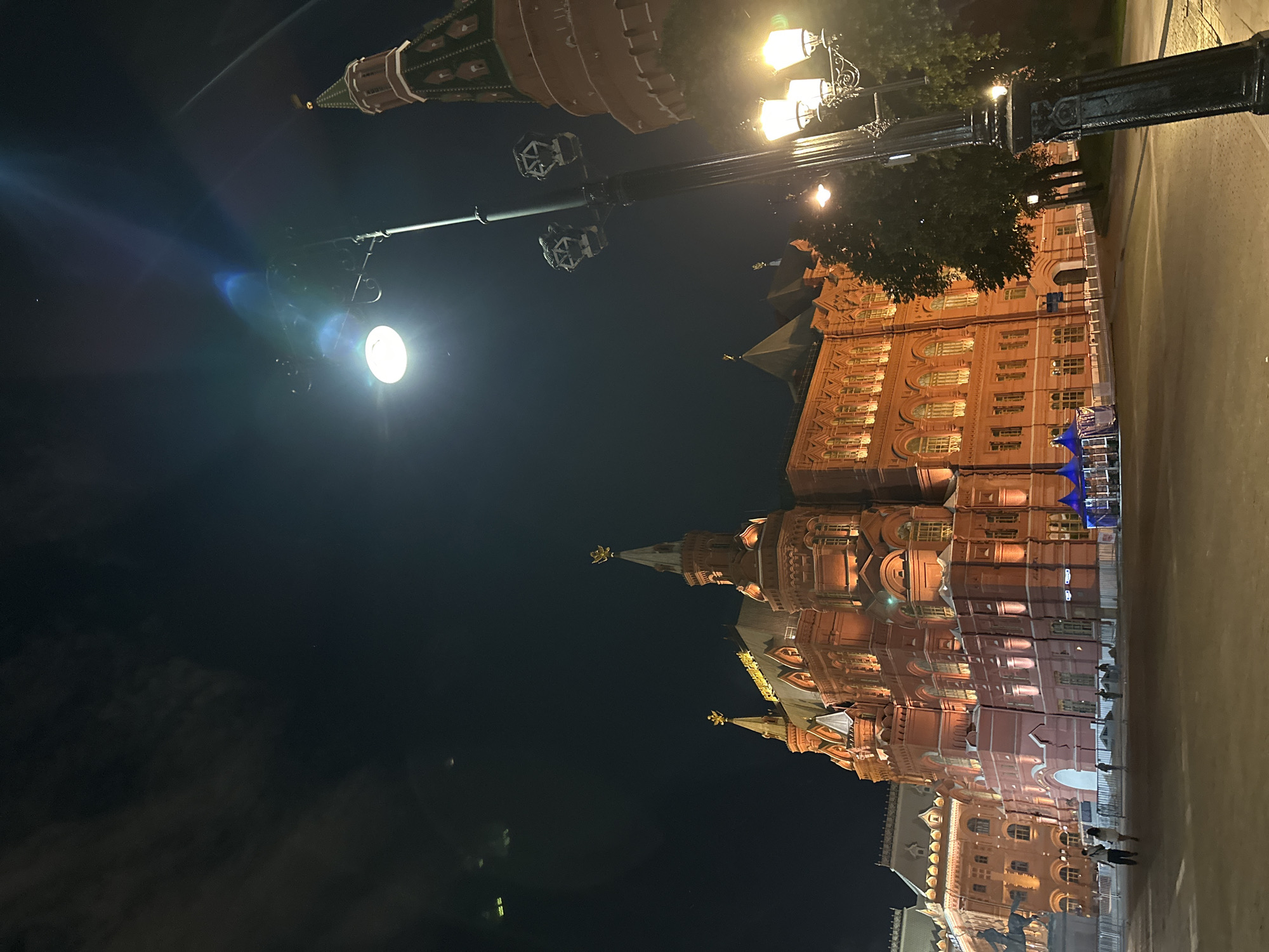The image depicts a nighttime scene in Moscow, Russia, featuring prominent historical landmarks under artificial illumination. The primary subject is the State Historical Museum, a large, ornate red-brick building with distinctive spires, pointed roofs, and intricate architectural details. Its facade is brightly lit by warm, golden lights, highlighting its historical grandeur.
To the left, a section of a Kremlin tower, possibly Nikolskaya Tower, is visible against the dark sky, characterized by its traditional green-capped spire and crenellated base. The sky is dark blue-black, indicating deep night.
The foreground and midground are illuminated by several streetlights. One bright, modern-looking streetlight is prominent on the left, casting a strong glow and causing a lens flare. Further into the scene, more traditional, ornate street lamps with multiple bulbs cast warm light onto the paved ground and a section of trees.
A few indistinct figures, appearing to be people, are visible near the base of the State Historical Museum, suggesting some pedestrian activity despite the late hour. The ground in the foreground appears to be paved, resembling a street or pedestrian area.
Notable details include the clear night sky, the stark contrast between the brightly lit buildings and the dark background, and the elaborate historical architecture. Golden decorative elements are visible on the roofline of the State Historical Museum, though no specific text is clearly legible. The overall impression is one of a majestic, historic city square at night.