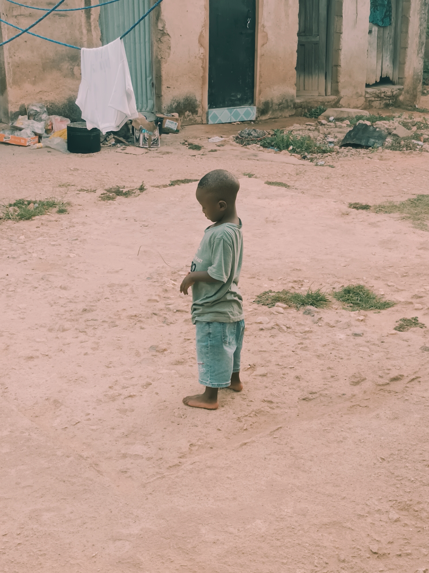 The photograph presents a full-body, side-view shot of a young, Black child standing barefoot on a dusty, uneven ground surface. The child is wearing a light gray t-shirt and blue denim shorts. Their head is shaved close to the scalp. They stand near a building or structure with worn, textured walls and two visible entrances. One entrance is a dark, rectangular door, while the other is a dilapidated wooden door. Debris and objects are scattered near the base of the building, including what appear to be plastic bags, a black container, and other assorted items. Clothes hang on a blue line strung from the building. Patches of green vegetation dot the ground. The location is identified as Bukuru, Nigeria.