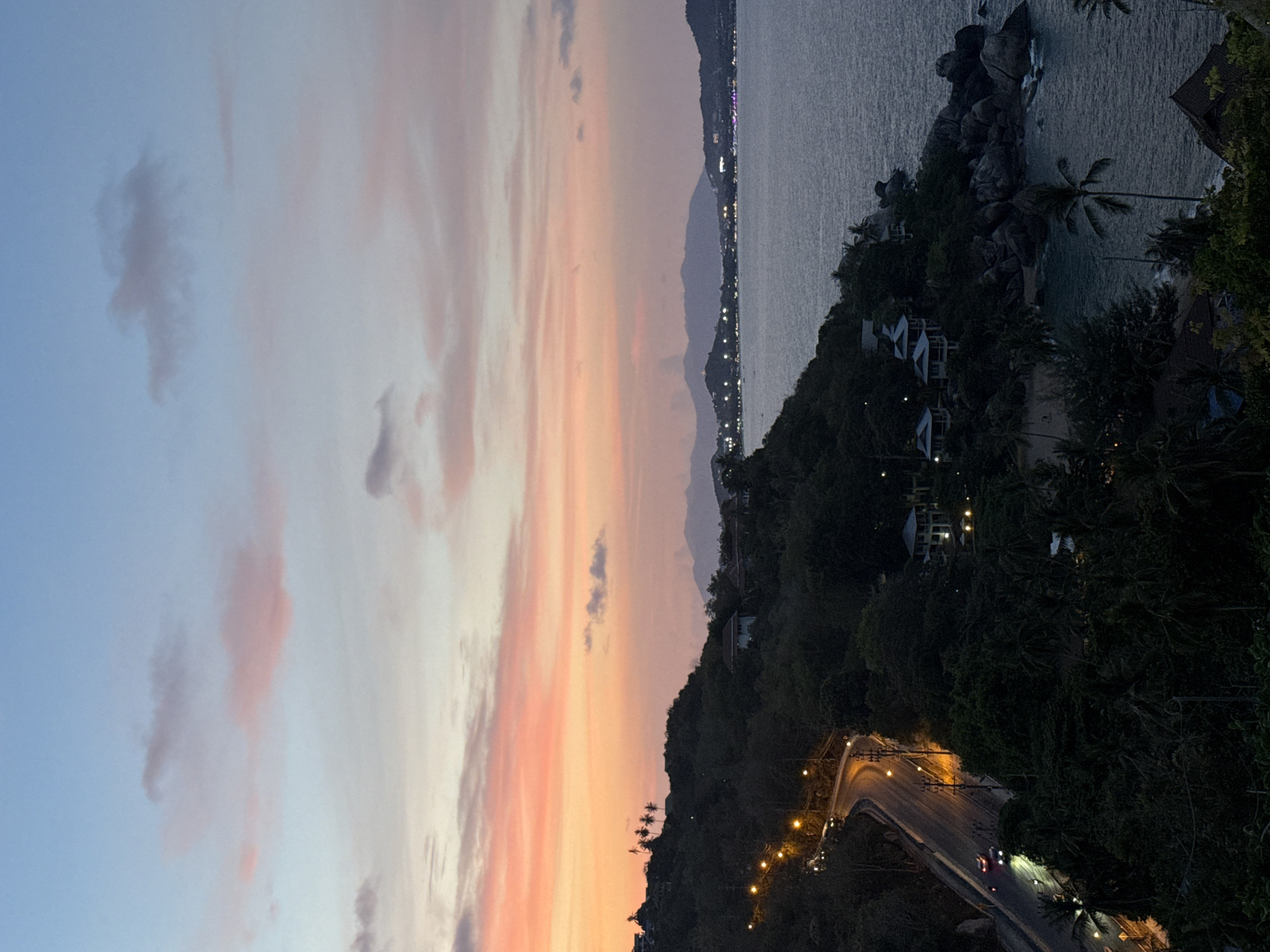 Baan Chaweng Noi, Thailand at dusk, exhibiting a vibrant sunset. The sky transitions from light blue to shades of pink and orange. A road illuminated by streetlights winds along the coastline, with moving vehicles visible. Dense vegetation covers the hillside leading to the coast. The sea, reflecting the remaining daylight, meets a rocky shoreline dotted with palm trees and buildings. In the distance, city lights run along the coastline.