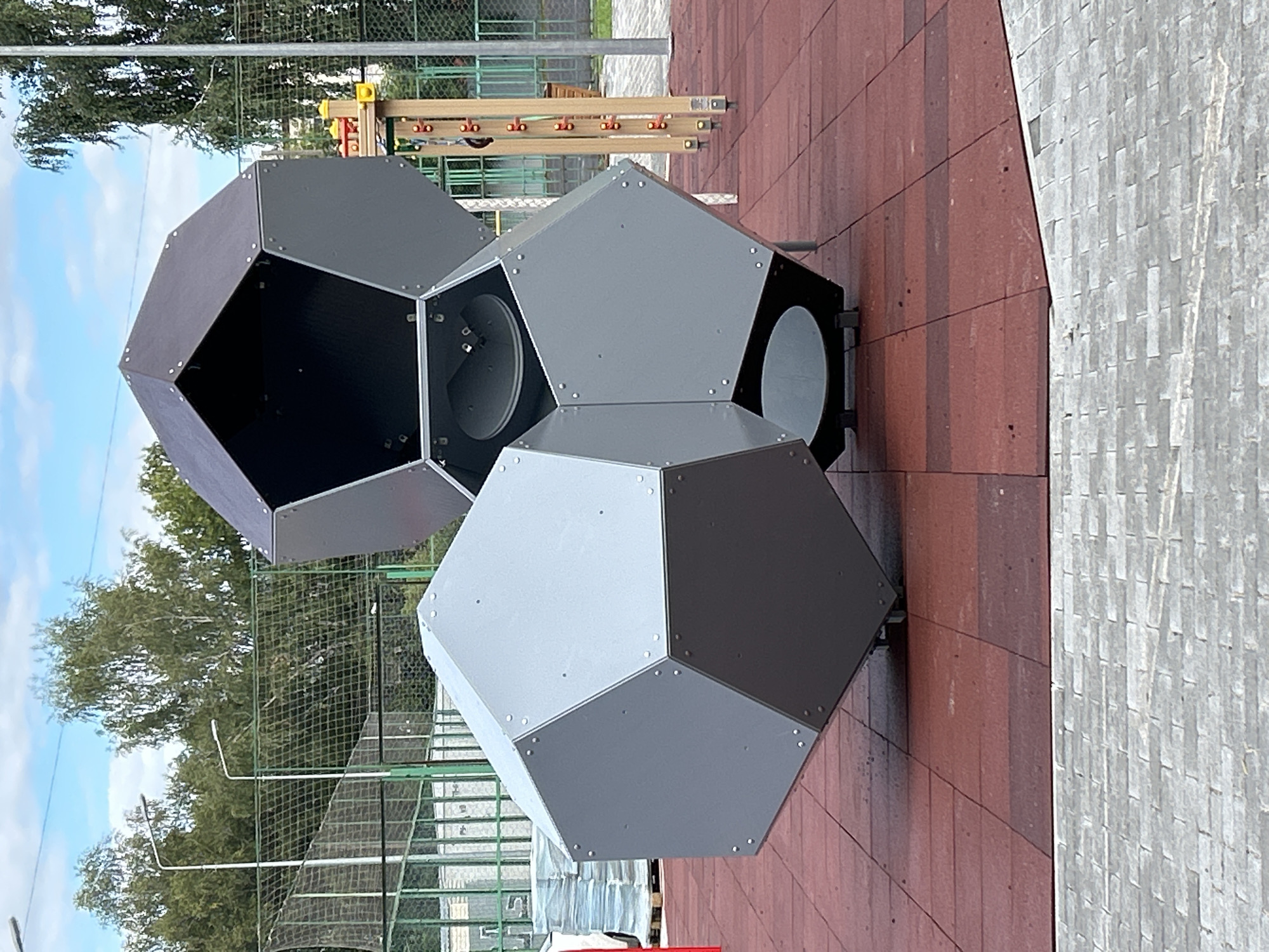 The image depicts an outdoor playground scene in Tomsk, Russia. The primary focus is on two large, geodesic dome-like structures made of grey and dark grey panels, assembled with visible rivets. These structures appear to be playground equipment, with openings that suggest they might be enclosed play areas or tunnels. One structure is angled, with its base resting on a red rubberized safety surface. The other, partially obscured, is positioned behind and above the first, its dark top section angled towards the viewer.
The playground surface is a mosaic of red rubber tiles, transitioning to a lighter, grey brick-like pavement in the foreground. In the background, a green metal fence encloses a sports field with a net. Beyond the fence, trees with green foliage are visible under a partly cloudy sky. Streetlights are present along the fence line. Some building structures and stacks of materials on pallets are also visible in the distance, to the left of the playground. The overall impression is of a newly constructed or renovated public recreational space.