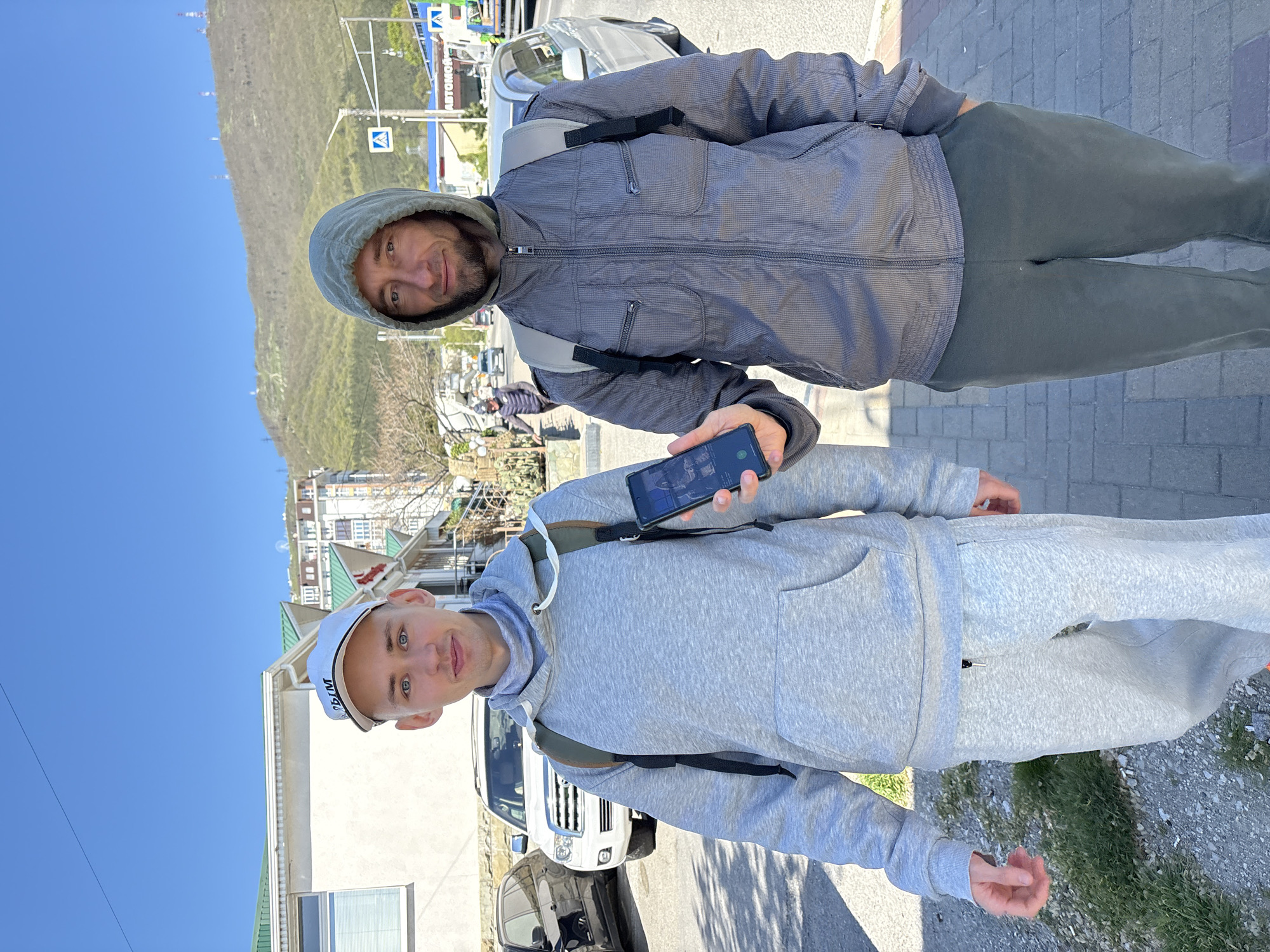 Two men are captured in an outdoor, full-body portrait on a sunny day, likely in Gelendzhik, Russia, given the provided context. The image is taken along a paved street or sidewalk, with a large, tree-covered mountain visible in the background under a clear blue sky.
The man in the foreground, younger, stands closer to the camera. He wears a light grey hooded sweatshirt, matching light grey sweatpants, and a white baseball cap with the Russian word "ДЫМ" (DYM, meaning 'smoke') visible on the front. He looks directly at the camera with a neutral expression, and a dark-strapped backpack is visible over his shoulders. His left hand is slightly open, while his right arm is mostly obscured.
Behind him and slightly to the right, an older man is smiling towards the camera. He wears a dark grey, striped hooded jacket, dark green pants, and a light-colored backpack. In his right hand, he holds a black smartphone, its screen displaying an image, possibly a map or another photograph.
The setting features urban elements mixed with natural scenery. To the left, a steep, green hillside or mountain dominates the background. To the right, various buildings, including a white house with green roofs, are visible. Several vehicles, including a white SUV, are parked along the street. The ground is paved with grey interlocking bricks, and some sparse vegetation like grass and small trees are present. A blue square pedestrian crossing sign is clearly visible in the mid-background, and part of another sign with text resembling "AUTOMOBIL" can be seen further back. The bright sunlight suggests it is daytime.