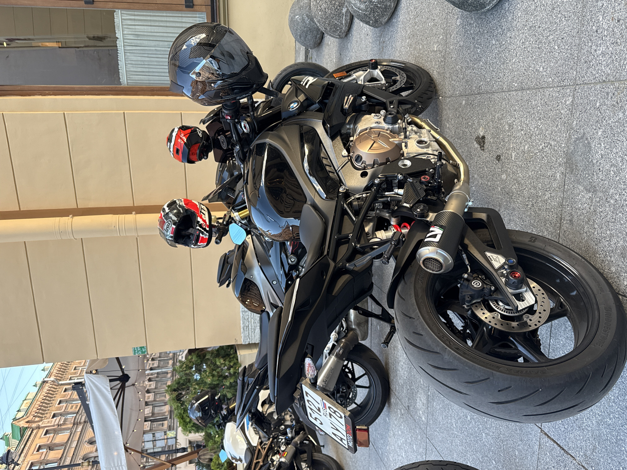 The image shows a row of motorcycles parked on a paved sidewalk in front of a building. The motorcycles are black and red, with helmets on top of them. One motorcycle has a license plate that says "5127 AX 78 RUSSIA". In the background, there is a building with windows and a white awning. The overall scene appears to be a street in Saint Petersburg, Russia, given the license plate and architectural style. The lighting suggests it is daytime.