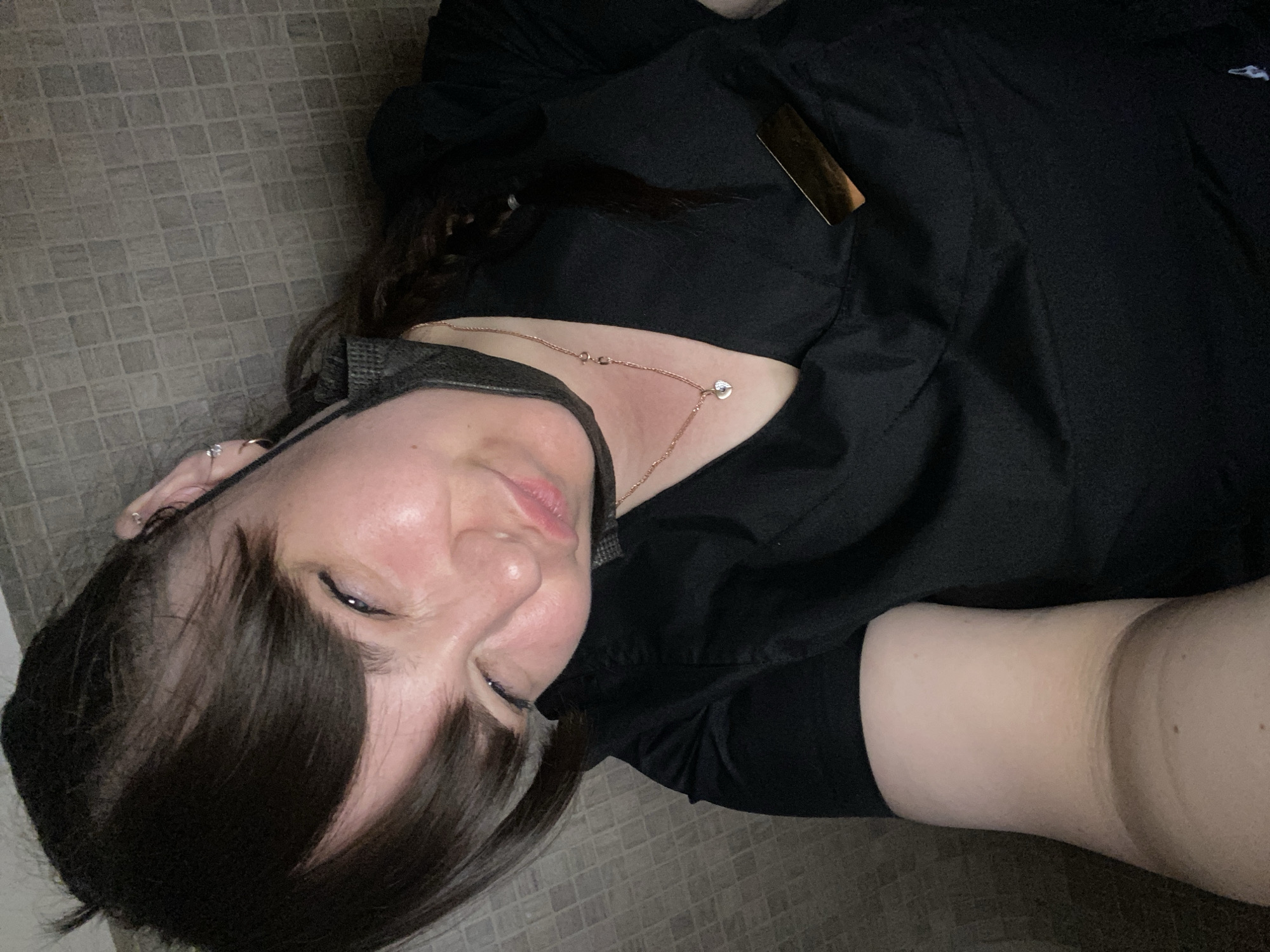 A woman with dark hair is lying on a light brown tiled floor. She is wearing a black mask and a black top. A gold chain necklace is visible around her neck. The location is unknown and cannot be determined from the image.