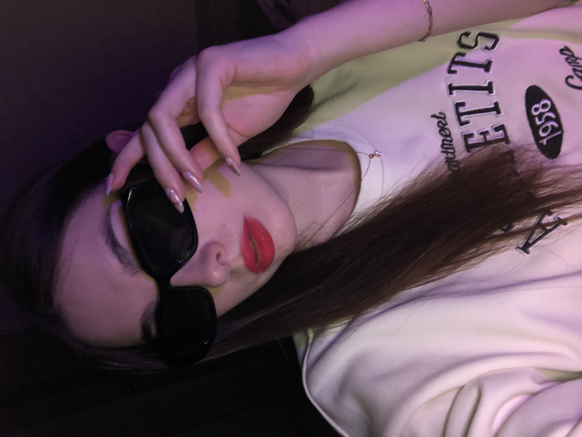 A woman with long, brown hair is wearing large black sunglasses and bright red lipstick. She is wearing a light-colored sweatshirt with black lettering that includes "1958." She is wearing a thin gold bracelet and has long, pointed nails with designs. She is holding the sunglasses with her left hand. The lighting is somewhat dim and appears to have a purple hue. It is impossible to determine her location from the image.