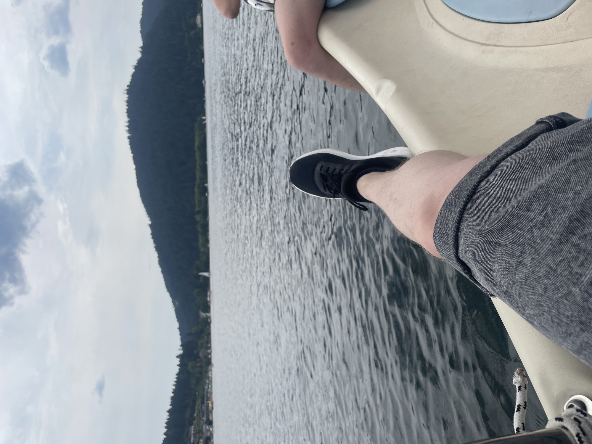The image shows a person's legs and feet propped up on the edge of a boat on a lake. The person is wearing gray shorts and black sneakers. The lake is calm, with ripples on the surface. In the background, there is a tree-covered mountain and a cloudy sky. The scene is set in the city of Gérardmer, France, known for its lake.