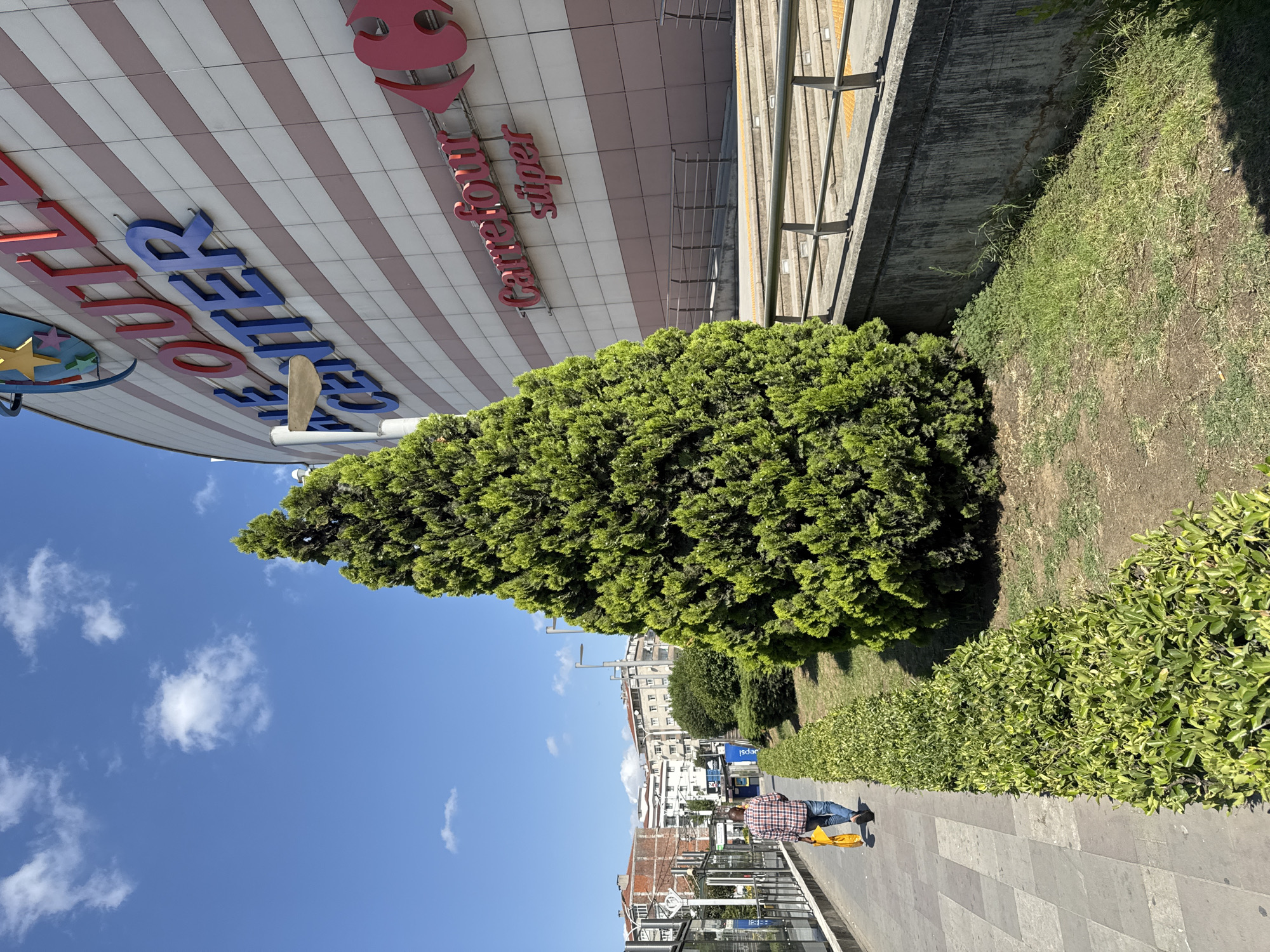 A man walks away from the camera on a sidewalk next to a large, modern shopping center in Bahçelievler, Turkey. The shopping center has the words "THE OUTLET CENTER" in large blue and red letters on its facade, along with a Carrefour logo. A tall, green evergreen tree stands in front of the shopping center, obscuring a portion of it. The sidewalk is paved with large, gray square tiles and bordered by a neatly trimmed hedge. In the background, residential buildings and streetlights are visible. The sky is a clear blue with a few scattered white clouds. The man is wearing a red and blue plaid shirt, blue jeans, and is carrying a bright yellow bag. The overall scene depicts an ordinary day in a commercial area of the city.