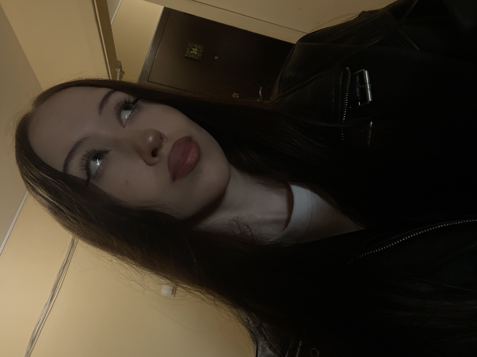 A young woman with long, dark hair and light-colored eyes is the sole subject in this close-up, low-angle, indoor portrait. She is wearing a black leather jacket over a white garment. Her gaze is directed upwards and to her left, with her mouth slightly open and her tongue extended, suggesting a playful or flirtatious expression.
The background is dimly lit, with a warm yellow wall visible on the left. To the right, a dark wooden door or cabinet is present, with a small, square plaque displaying the number "34" in a gilded font. The lighting is dim, casting shadows and highlighting the woman's features, particularly her eyes and lips. The overall mood is intimate and somewhat mysterious due to the low light conditions. No specific time of day or weather conditions can be determined from the image. The visible text is limited to the number "34" on the door. The setting appears to be a domestic interior.