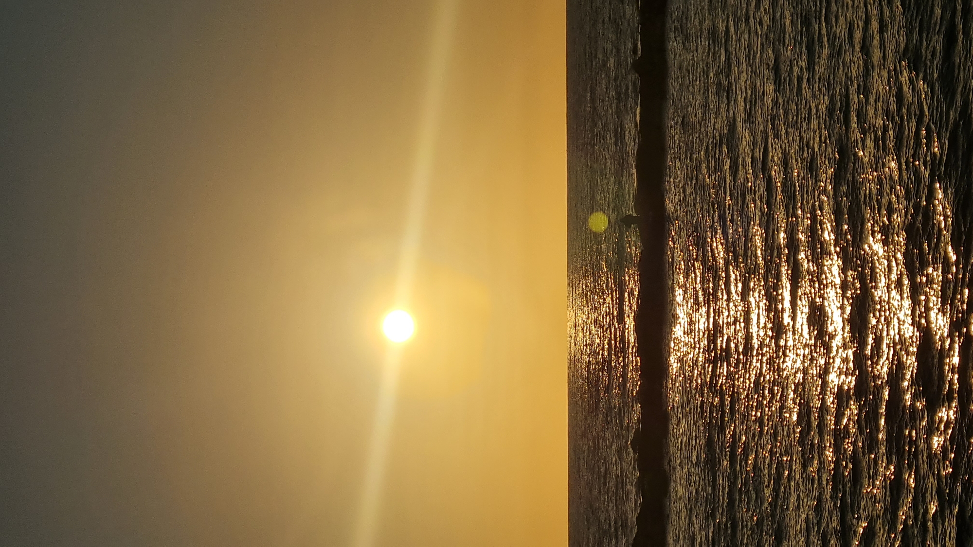 The image captures a sunset scene in Nahariyya, Israel. The sun is positioned to the left, its bright light dominating the sky, which is bathed in a golden hue. To the right, the sea reflects the sunlight, creating a shimmering effect on the water's surface. A dark, vertical object, possibly a pier or structure, divides the sky and the sea.