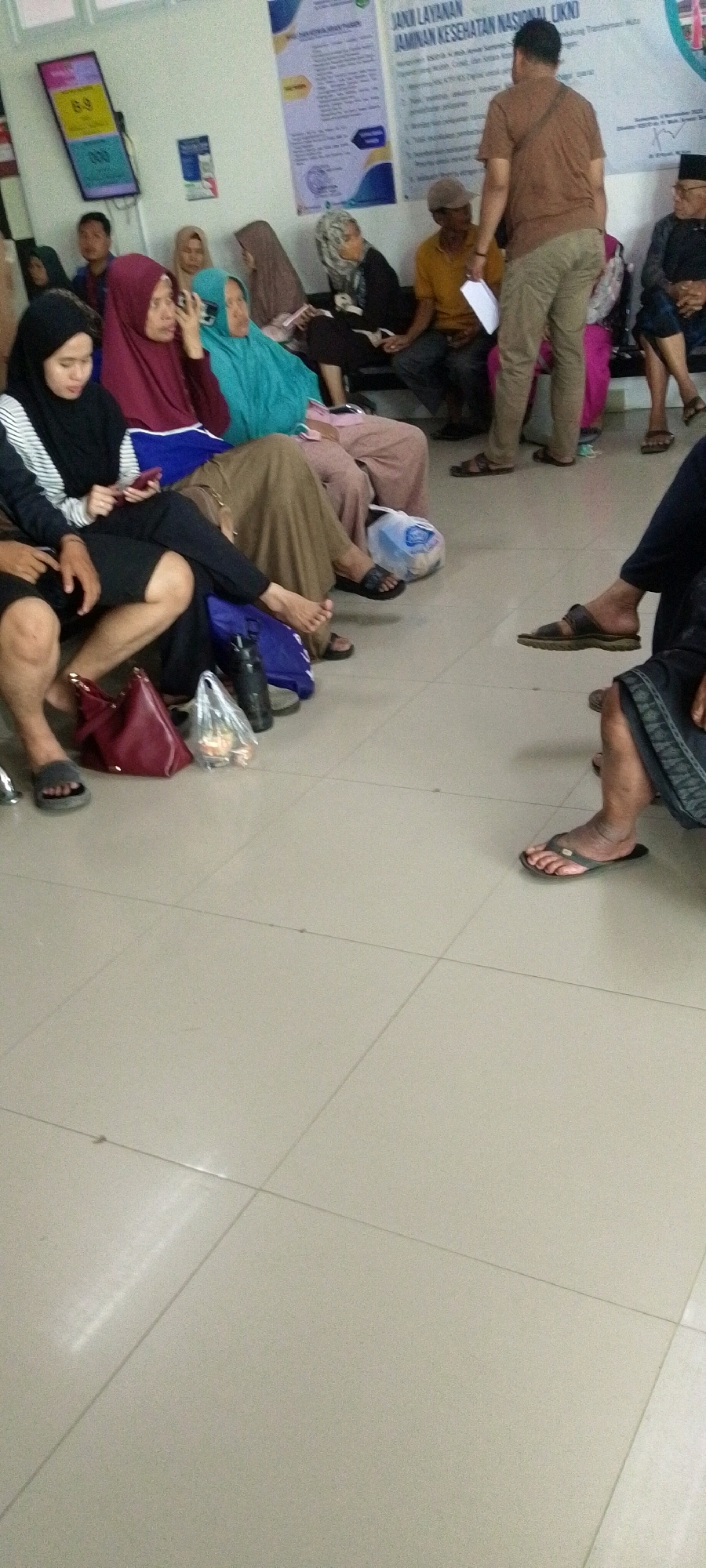 The image shows a waiting area, likely at a public health facility, filled with people sitting and standing. Several individuals are wearing headscarves, common attire in Indonesia. The room has a tiled floor and a wall with posters, one of which displays information in Indonesian. A digital display shows numbers, possibly for queue management. Bags and personal belongings are scattered around the seating area. The scene depicts a typical day in a public waiting room, with people patiently awaiting their turn. It is likely located in Sumenep, a regency in East Java, Indonesia, given the language on the posters and the cultural context.