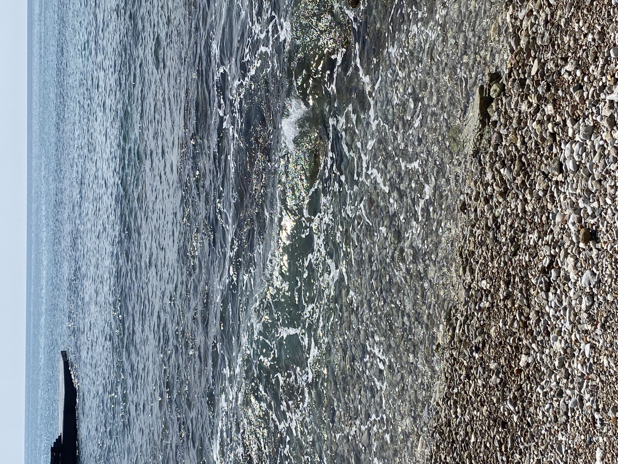The image depicts a coastline with water meeting a pebble beach. The water is textured with visible waves and areas where the pebbles beneath are visible. The beach consists of densely packed, small, rounded pebbles. A wooden structure or remnant of a pier is seen extending into the water on the left. The sky is overcast. The location is likely near Budva, Montenegro, given the typical coastline.
