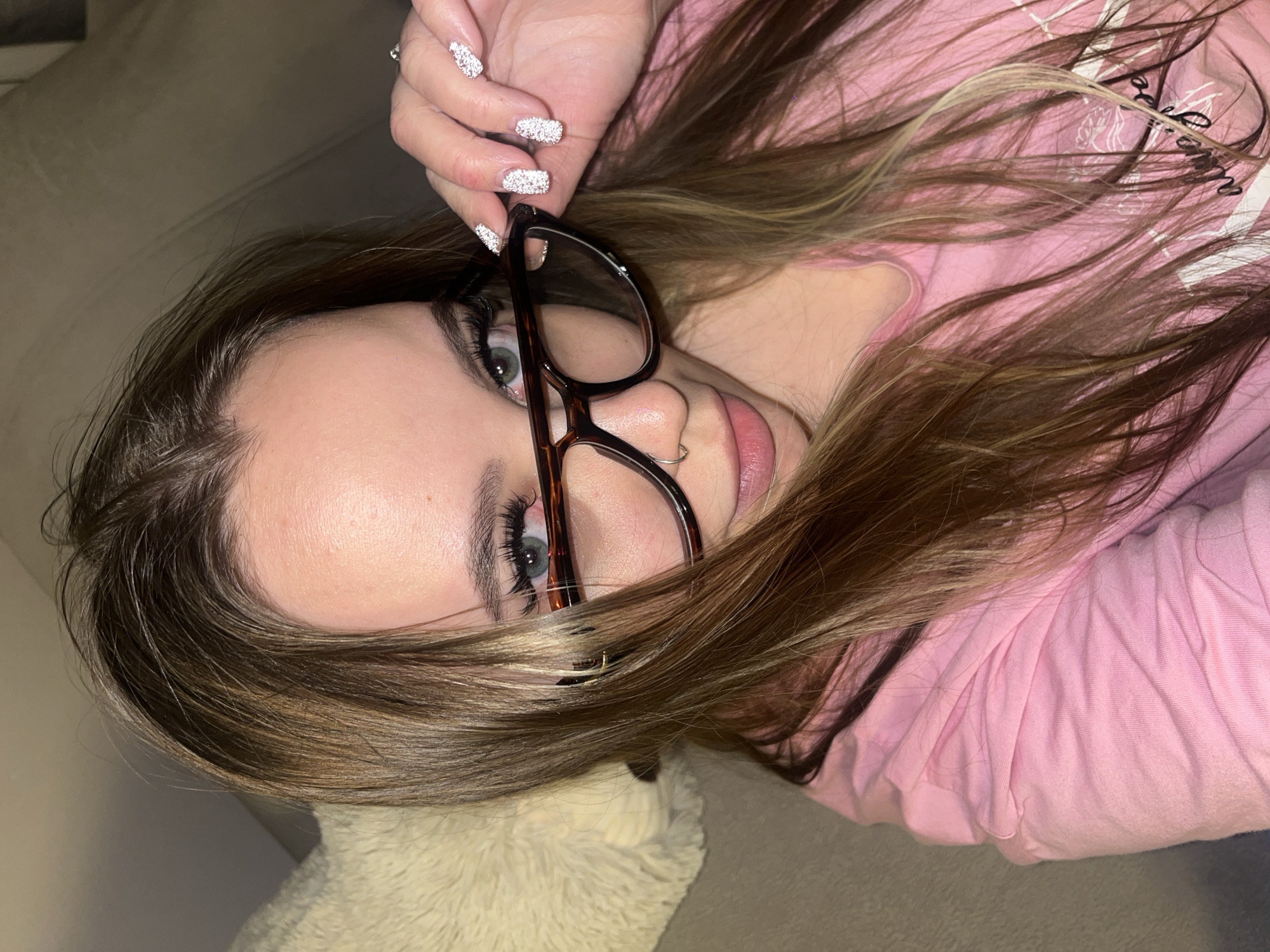 A young woman is lying down, turned slightly towards the viewer. She has light brown hair with blonde highlights and is wearing glasses. She is wearing a pink shirt. Her fingernails are decorated with sparkly embellishments. Part of what appears to be a light-colored cushion is visible near her head. The location is unknown and I cannot give a specific location in Krasnodar, Russia.