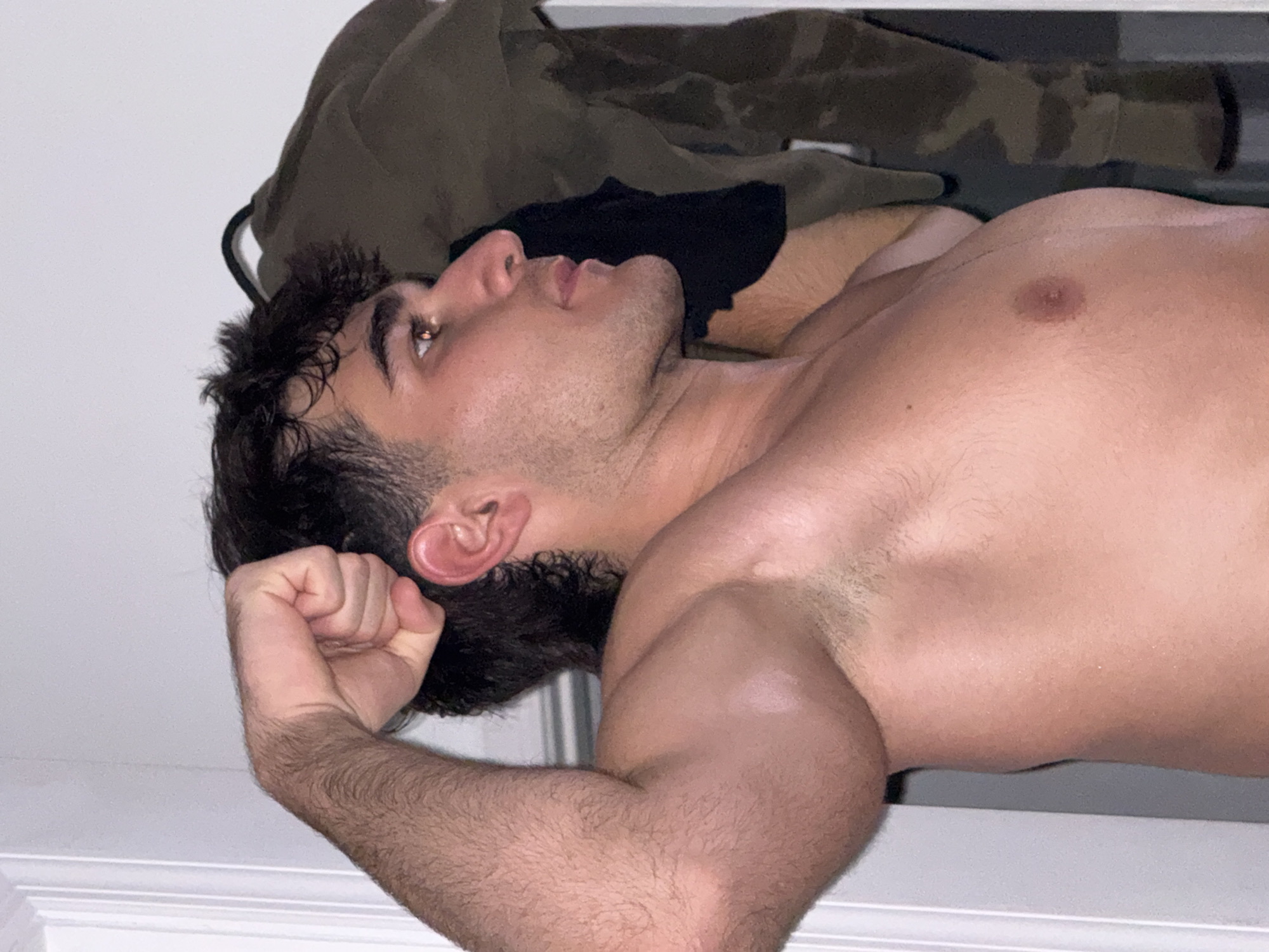 A young adult male, shirtless and with dark, curly hair, flexes his bicep while looking to the right. His left arm is raised, with his fist clenched. The background consists of a white door on the left and a rack of clothing on the right, including a green camouflage jacket. The image appears to be taken indoors, likely in a residential setting within Bellmore, United States of America.