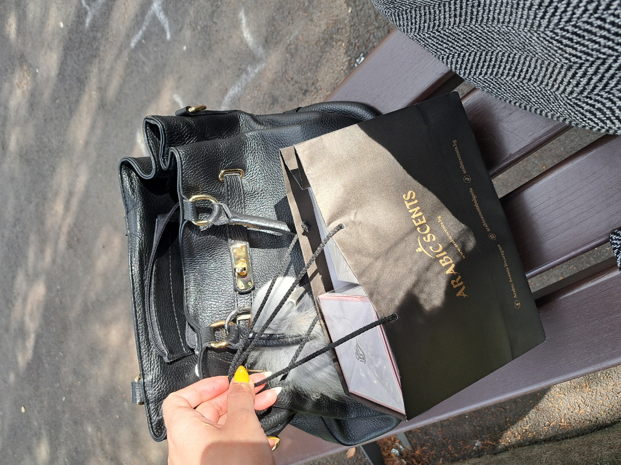 An overhead shot depicts an urban outdoor scene in Sofia, Bulgaria, likely a park or pedestrian area. A person is seated on a dark brown, slatted wooden bench, with a portion of their leg, covered in grey and white herringbone fabric, visible on the right. On the bench, to the left of the leg, rests a black pebbled leather handbag featuring gold-colored hardware, including a central buckle and D-rings. A grey faux fur accessory is attached to the handbag's strap. Adjacent to the handbag is a black paper shopping bag with "ARABIC SCENTS" in gold lettering and "www.arabicscents.bg" printed below. The bag also displays social media handles, including "Arabic Scents България". Inside the shopping bag, a light-colored rectangular box with a decorative emblem is partially visible. A human hand, positioned in the upper left, displays bright yellow nail polish and grasps one of the black string handles of the "ARABIC SCENTS" bag. The scene is brightly lit by sunlight, casting distinct shadows. The background consists of dark asphalt pavement with faded white markings.