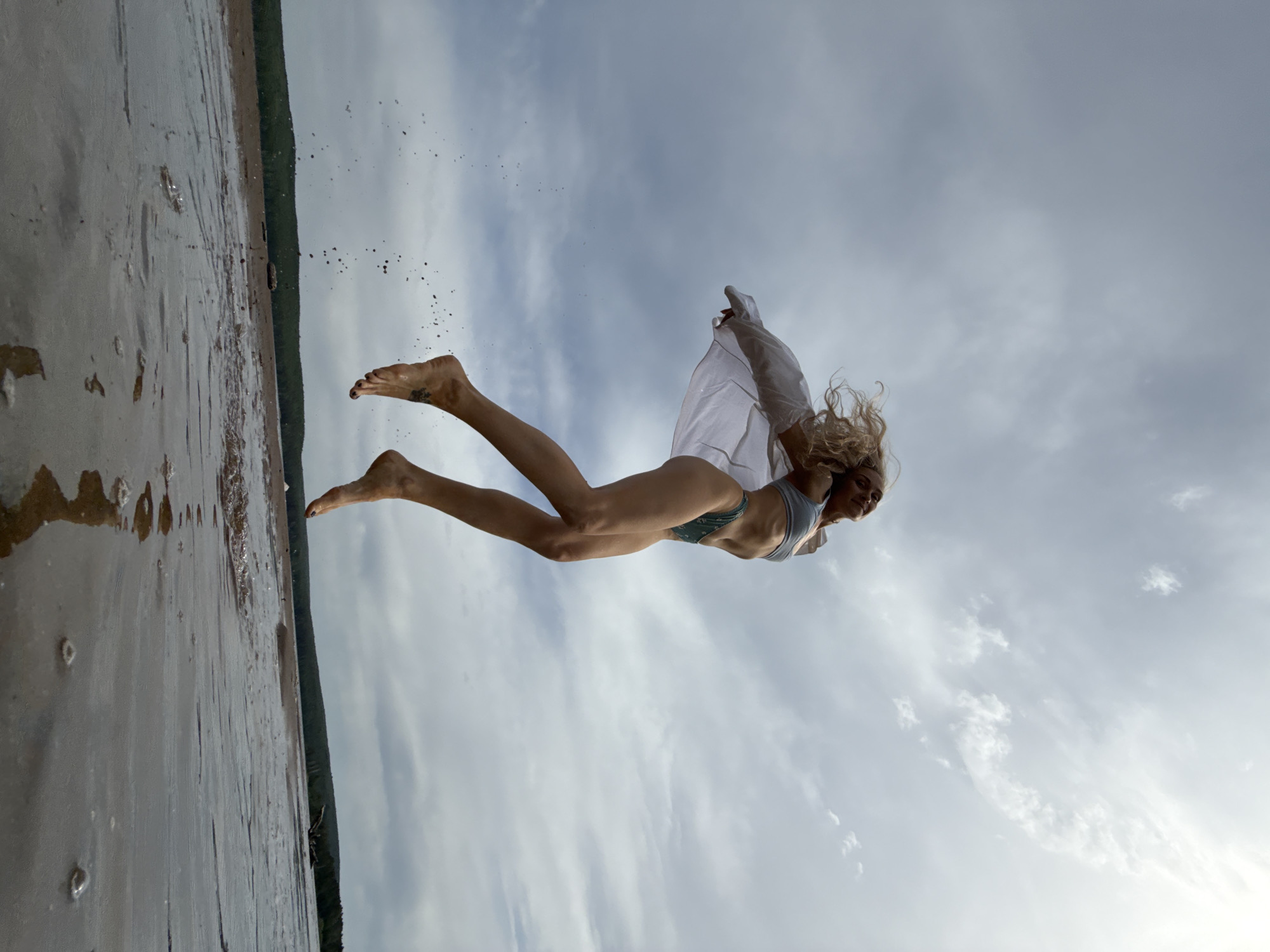A blonde woman in a gray bathing suit and a white cloth flies suspended in the air over what appears to be a beach or shoreline. The shoreline is along the left side of the frame, and the background is a cloudy sky. Dark water and wet sand line the shore. The exact location is Chaikovsky, Russia.