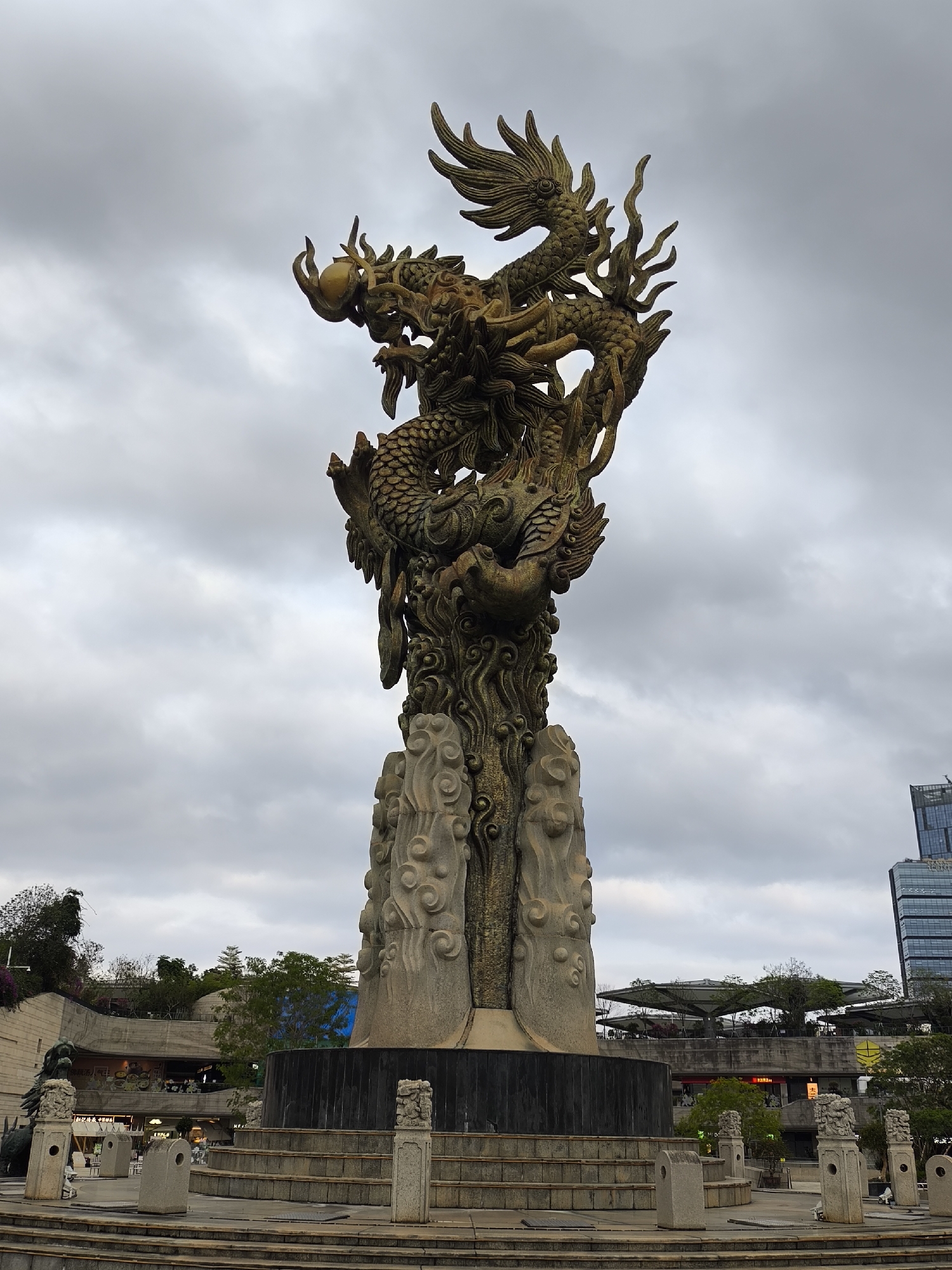 A bronze dragon statue sits atop a stone pillar in a public square in Longgang District, China. The dragon, with scales and wings, holds a ball in its claws. The stone pillar is decorated with cloud patterns. The statue is elevated on a stepped base with decorative stone posts. The square is surrounded by buildings, trees, and walkways. The sky is overcast. No people are visible in the immediate foreground.
